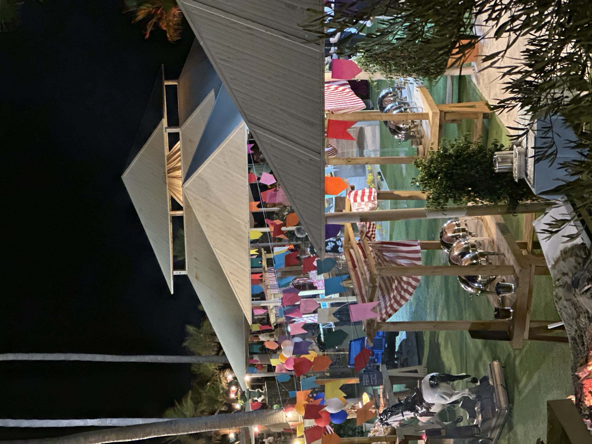 The image depicts an outdoor nighttime event, likely a celebration or party, set up on a grassy area. In the background, there is a large, multi-tiered white-roofed structure with visible wooden supports. Colorful triangular flags are strung across various parts of the scene, creating a festive atmosphere. Several wooden stalls or serving stations are set up, some displaying chafing dishes, indicating a food service area. In the foreground, to the left, a white rocking horse ride is visible. To the right, there are trees and shrubbery, with some olive branches in the immediate foreground. The ground appears to be artificial turf. There are a few people visible in the background, mostly indistinct. The lighting suggests the event is illuminated by artificial lights, creating a warm glow on the structures and food service areas. The presence of palm trees in the background hints at a warm climate, consistent with Estepona, Spain, a coastal town on the Costa del Sol known for its outdoor events and nightlife.
