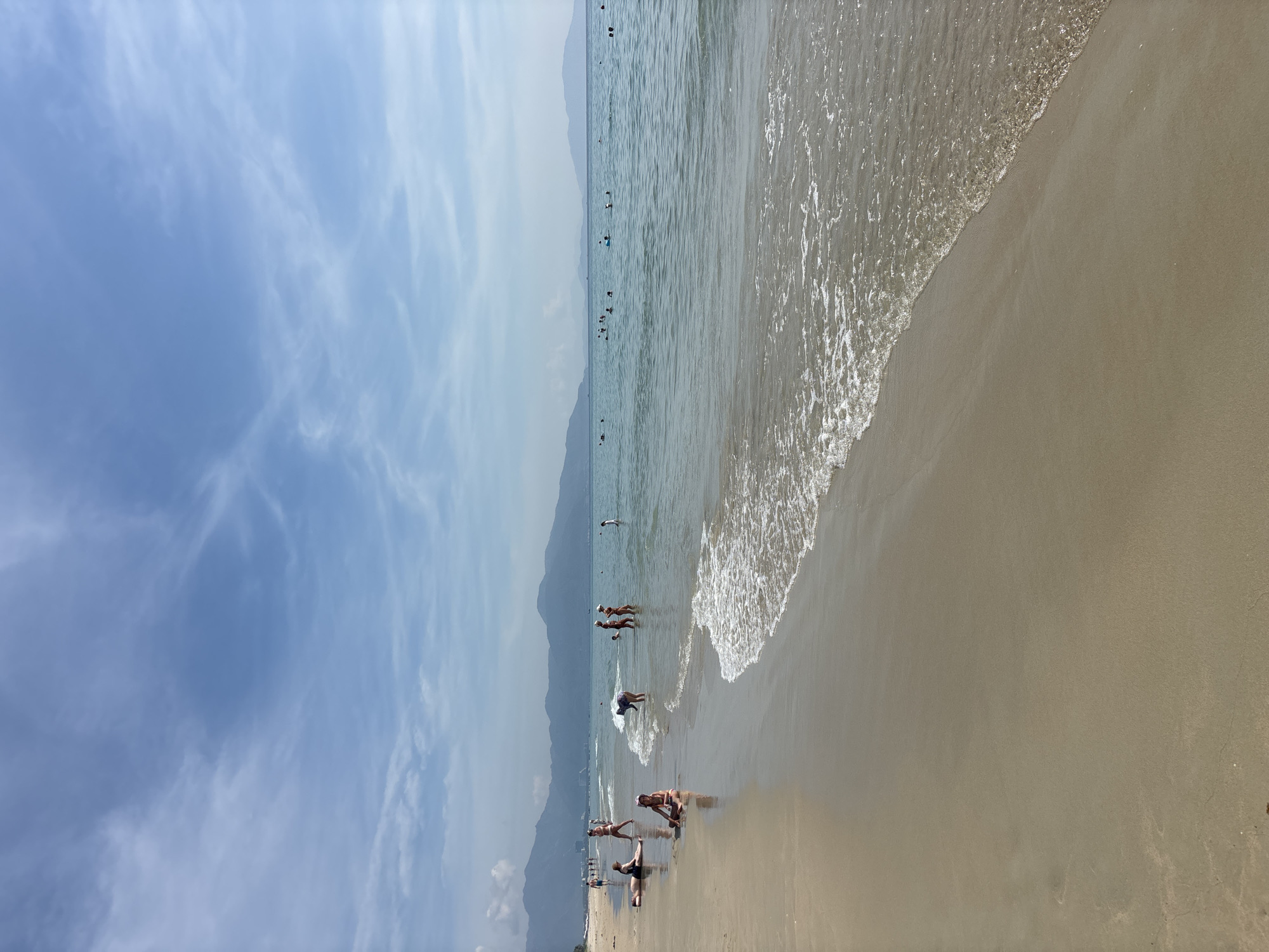 The image depicts a sunny day at a sandy beach with calm, clear water and distant mountains. The sky is a soft blue with wispy white clouds. Several people are visible in the shallow water and on the wet sand. Some are wading, while others appear to be enjoying a swim or playing in the water. A few individuals are relaxing on the beach, with one person in the foreground appearing to be doing some form of exercise. The tide is low, with gentle waves lapping at the shore. The light suggests it is daytime, likely afternoon, given the soft shadows and the brightness of the scene. The overall atmosphere is relaxed and leisurely, conveying a sense of a typical summer day spent at the seaside. There is no visible text in the image.