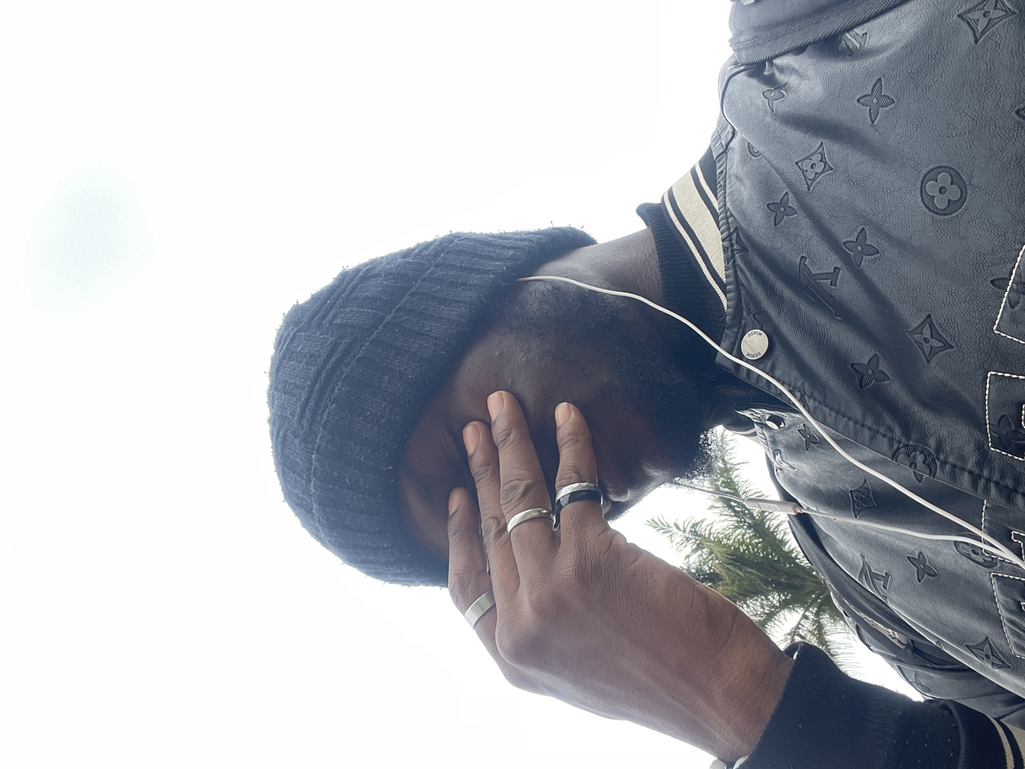 A person wearing a black beanie, a black patterned jacket, and white earbuds is captured from the chest up, outdoors against a bright, overcast sky. Their right hand is covering their eyes, and they are wearing several silver and black rings on their fingers. The jacket has a repeating pattern of Louis Vuitton-like symbols. In the background, a palm tree is visible, suggesting a warm climate. The image appears to be taken on a street or open area in Jos, Nigeria.