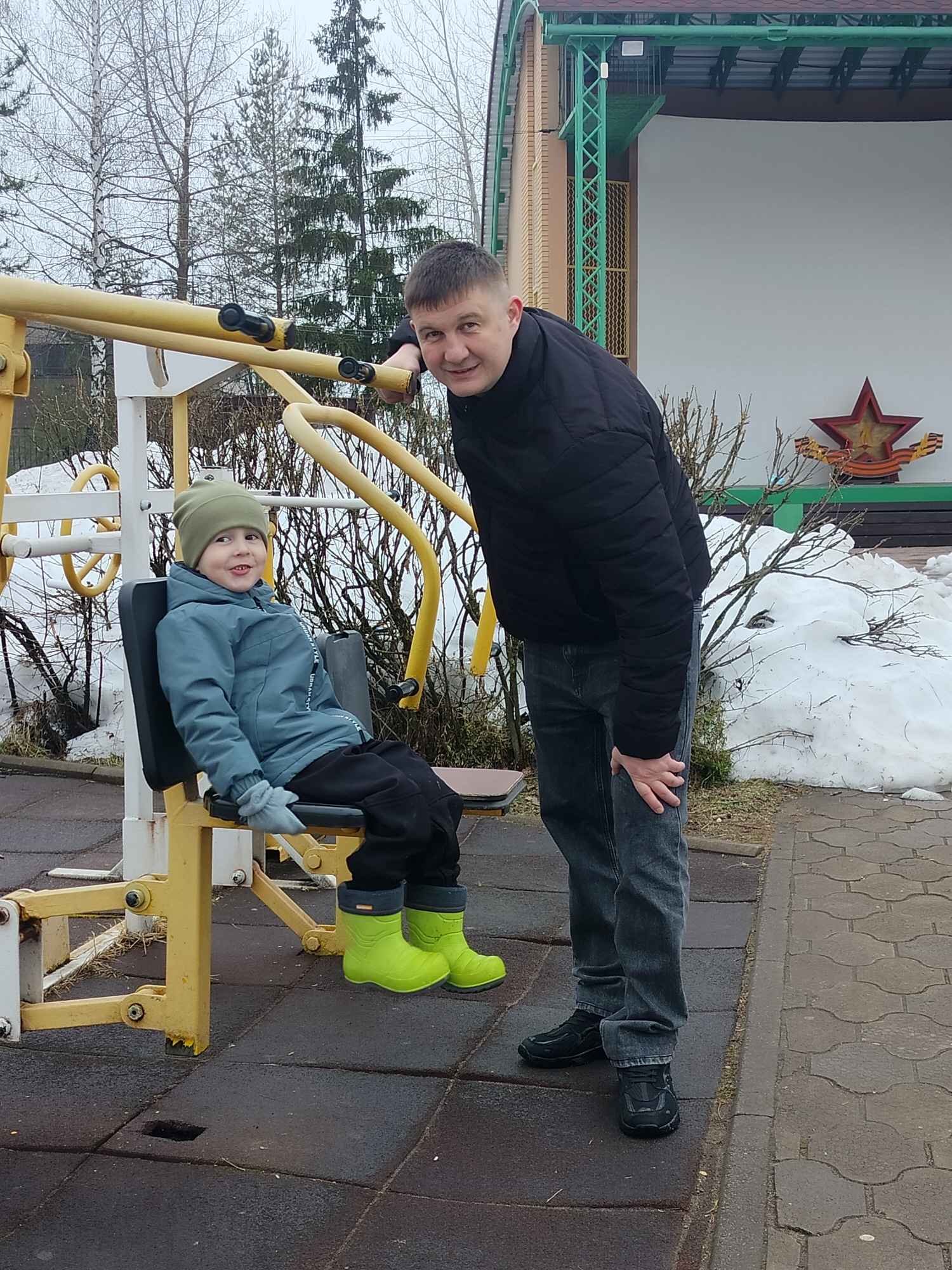 Two individuals are present in an outdoor setting. A young boy is seated on a yellow exercise machine. He is wearing a green hat, a blue jacket, black pants, and bright green boots. A man stands next to the machine, his hand resting on it, looking towards the camera. He is wearing a black jacket and gray jeans. Behind them is a building with a star emblem and residual snow. The location is Chaikovsky, Russia.