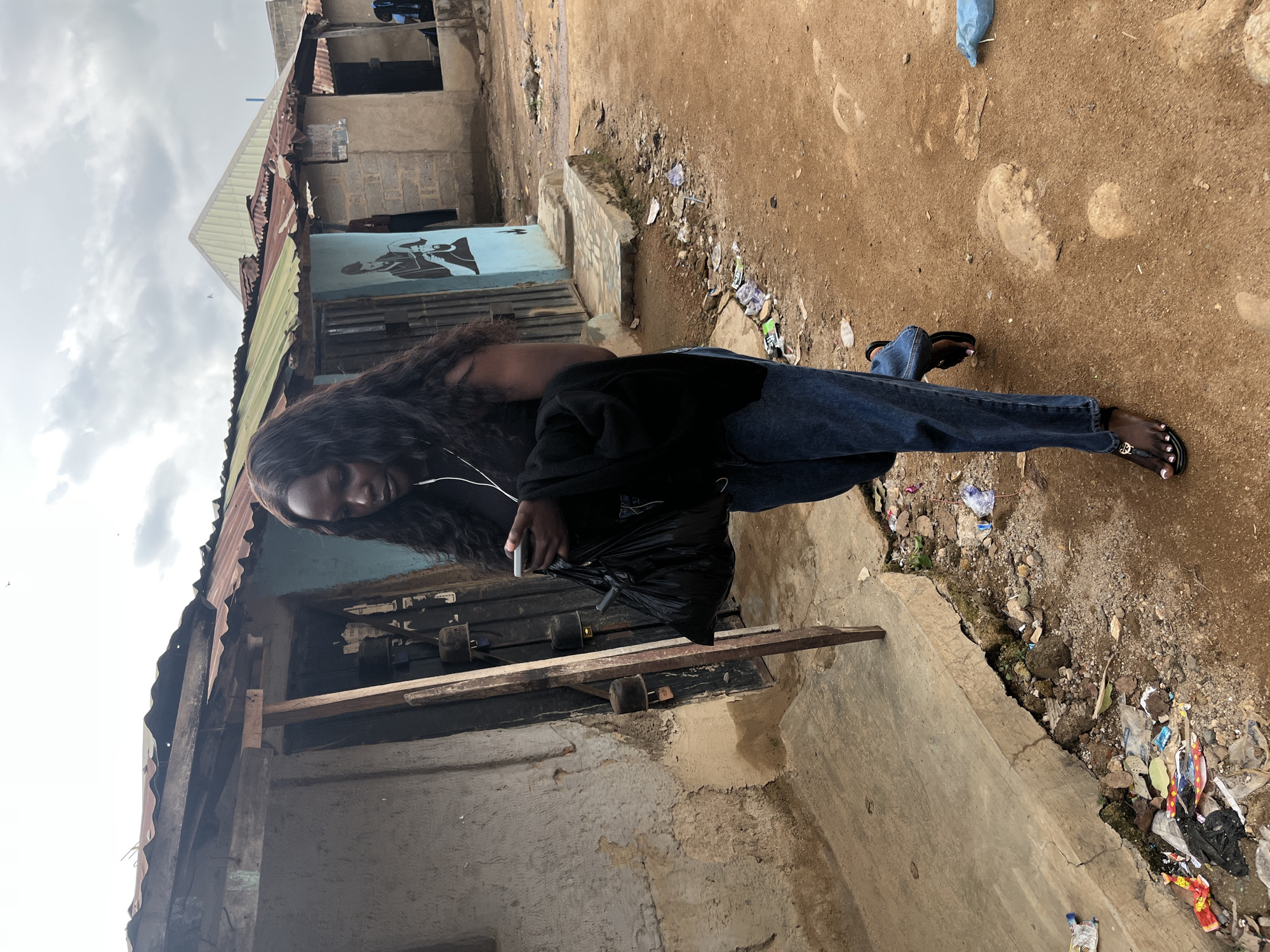 A young Black woman with long, dark, wavy hair is walking to the right in a dusty, unpaved street. She is wearing a black t-shirt, blue jeans, and black flip-flops. She is holding a black plastic bag in her left hand and a smartphone in her right hand, looking down at it. She has white earbuds in her ears. Behind her are several dilapidated buildings with corrugated metal roofs, some of which are rusted. The walls are a mixture of concrete and brick, with some adorned with faded paintings of people. The ground is covered in dirt and scattered with debris, including plastic bags and wrappers. The sky is overcast with some clouds visible. The scene appears to be in an impoverished urban area of Jos, Nigeria.