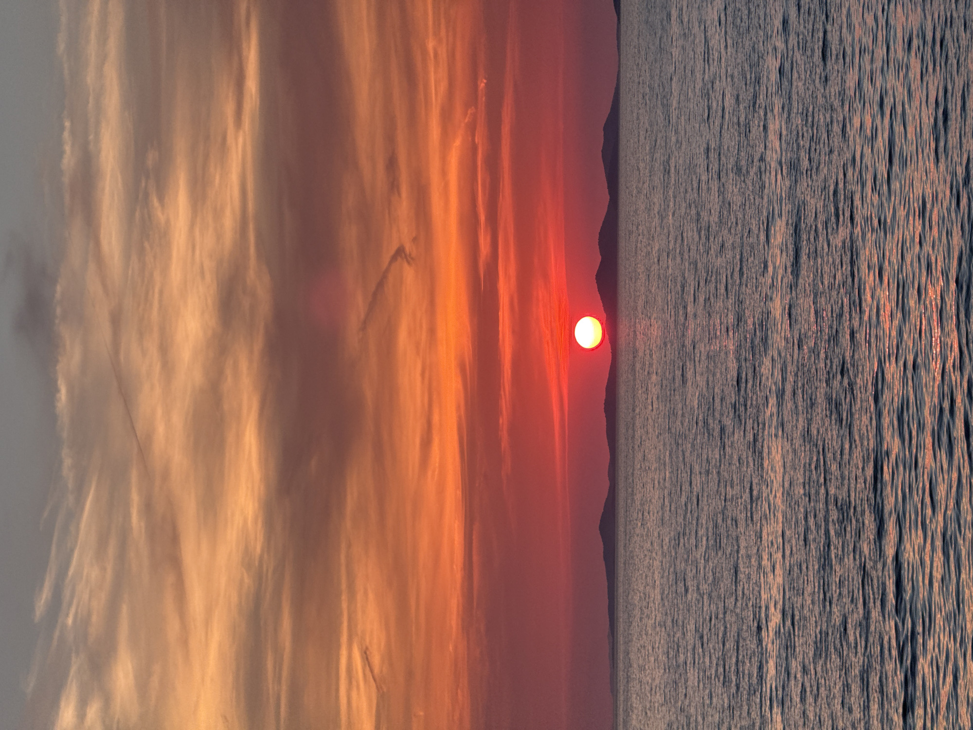 A vertical, eye-level shot captures a vibrant sunset over the Adriatic Sea, with the silhouette of islands in the distance, possibly near Primošten, Croatia. The sky is ablaze with fiery oranges and reds, transitioning to softer pinks and purples higher up, with wispy clouds streaked across. The sun, a brilliant white orb tinged with red, sits low on the horizon, casting a shimmering reflection on the rippling water below. The sea itself is a deep blue, with subtle hints of the sunset's colors reflecting on its surface. The overall impression is one of serene natural beauty at the end of the day.