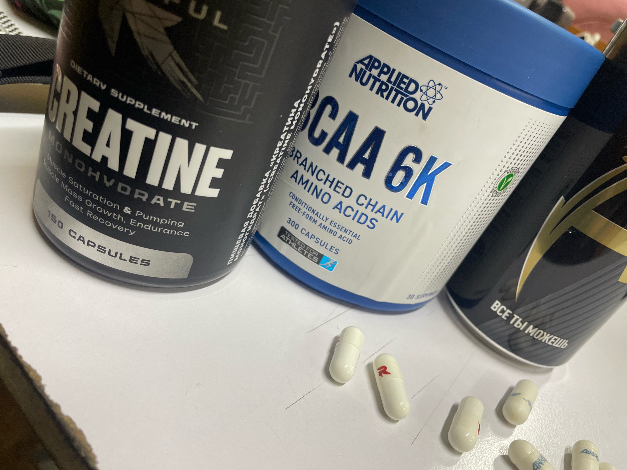 In an indoor setting, likely a table surface, several dietary supplement containers and capsules are arranged. Prominently featured is a black container labeled "CREATINE MONOHYDRATE" with text detailing its benefits like "Muscle Saturation & Pumping," "Muscle Mass Growth, Endurance," and "Fast Recovery." Next to it, a white and blue container displays "APPLIED NUTRITION BCAA 6K BRANCHED CHAIN AMINO ACIDS." A third, partially visible black container has text in Cyrillic, reading "ВСЕ ТЫ МОЖЕШЬ." Scattered on the surface are several white capsules, some with red markings. The lighting suggests an indoor environment, possibly with artificial light.