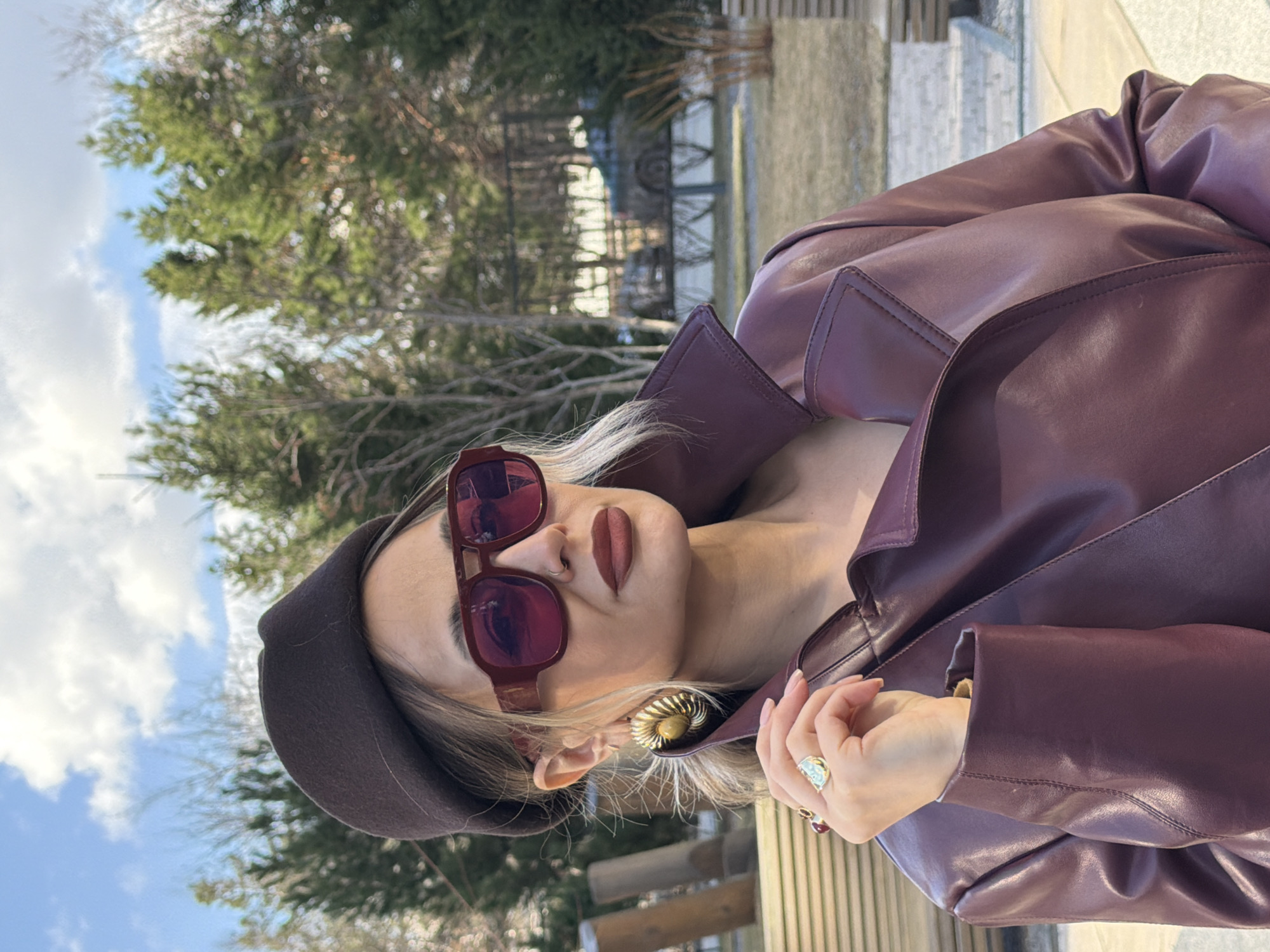 A head-and-shoulders portrait features a stylish woman posing outdoors in Krasnoyarsk, Russia. The image is presented sideways, showing the woman's head tilted back as she looks directly towards the camera. She wears large, reddish-brown square-framed sunglasses that obscure her eyes. Her lips are adorned with dark, matte reddish-brown lipstick, and a subtle ring piercing is visible on her nose. Light blonde hair peeks out from under a dark brown beret worn on her head.
The woman is dressed in a dark reddish-purple (burgundy or maroon) faux leather or leather jacket or coat, which features prominent lapels and stitching details. A large, intricate gold-toned statement earring with a spiral design is visible on her right ear. Her left hand, with manicured nails and a distinctive ring with a light-colored stone on her ring finger, is positioned near her chest. Her right hand is partially visible, holding a lapel of her jacket.
The background is softly blurred but reveals an outdoor setting under a bright, partly cloudy sky with patches of blue. Evergreen trees and bare deciduous trees, suggesting late winter or early spring, are visible. Faint architectural elements or fencing can be seen further in the background, hinting at a park or urban outdoor location. The bright daylight highlights the textures of her attire and accessories. There is no visible text in the image.