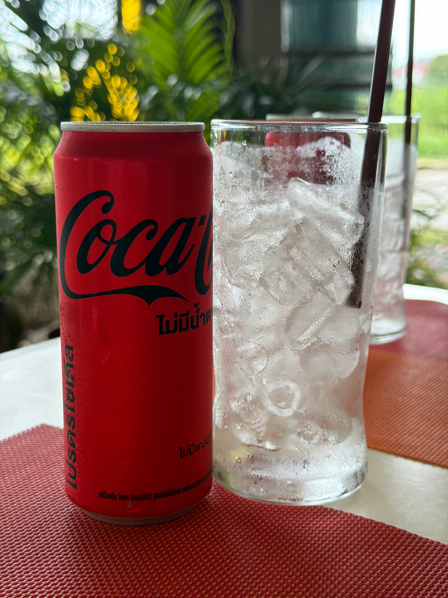 The image shows a close-up view of a red Coca-Cola can and a glass filled with ice and a dark straw, set on a textured red placemat. The Coca-Cola can is on the left, featuring the brand's iconic logo in black. Below the logo, there is text in Thai script. To the right of the can, a clear glass is filled with ice cubes and a dark straw. Condensation is visible on the outside of the glass, indicating it is cold. In the background, out of focus, are lush green foliage and yellow flowers, suggesting an outdoor or semi-outdoor setting, possibly a restaurant or cafe. Another glass with ice and a straw is partially visible behind the primary glass, suggesting a second drink. The overall scene appears to be taken in Hua Hin City Municipality, Thailand, based on the presence of Thai script on the Coca-Cola can. The lighting suggests it is daytime.