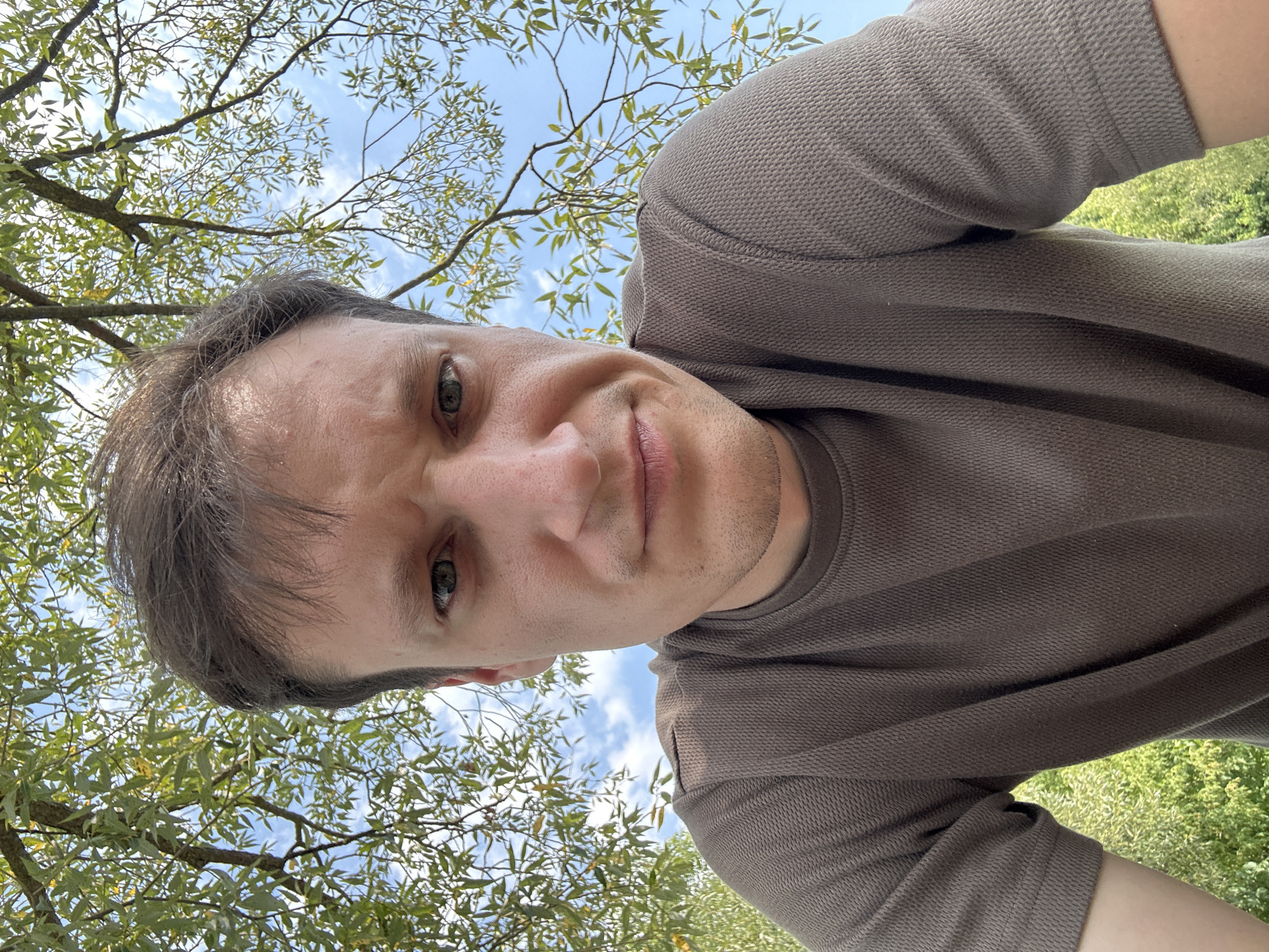 The image is a low-angle, close-up selfie of a man taken outdoors. He is looking directly at the camera with a neutral to slightly concerned expression. His hair is brown and medium length, with some strands falling forward onto his forehead. He has a fair complexion and light-colored eyes. He is wearing a textured, short-sleeved brown t-shirt.
The background consists of lush green foliage from tree branches with light green leaves, and a bright blue sky with scattered white clouds. The dappled sunlight suggests it is daytime. The presence of trees and greenery indicates an outdoor, possibly park or garden setting within the city of Makiv, Ukraine. There are no other people or specific man-made objects visible in the immediate vicinity that would provide further context about the location. The framing of the shot focuses primarily on the man's face and upper torso, with the natural environment serving as a backdrop.