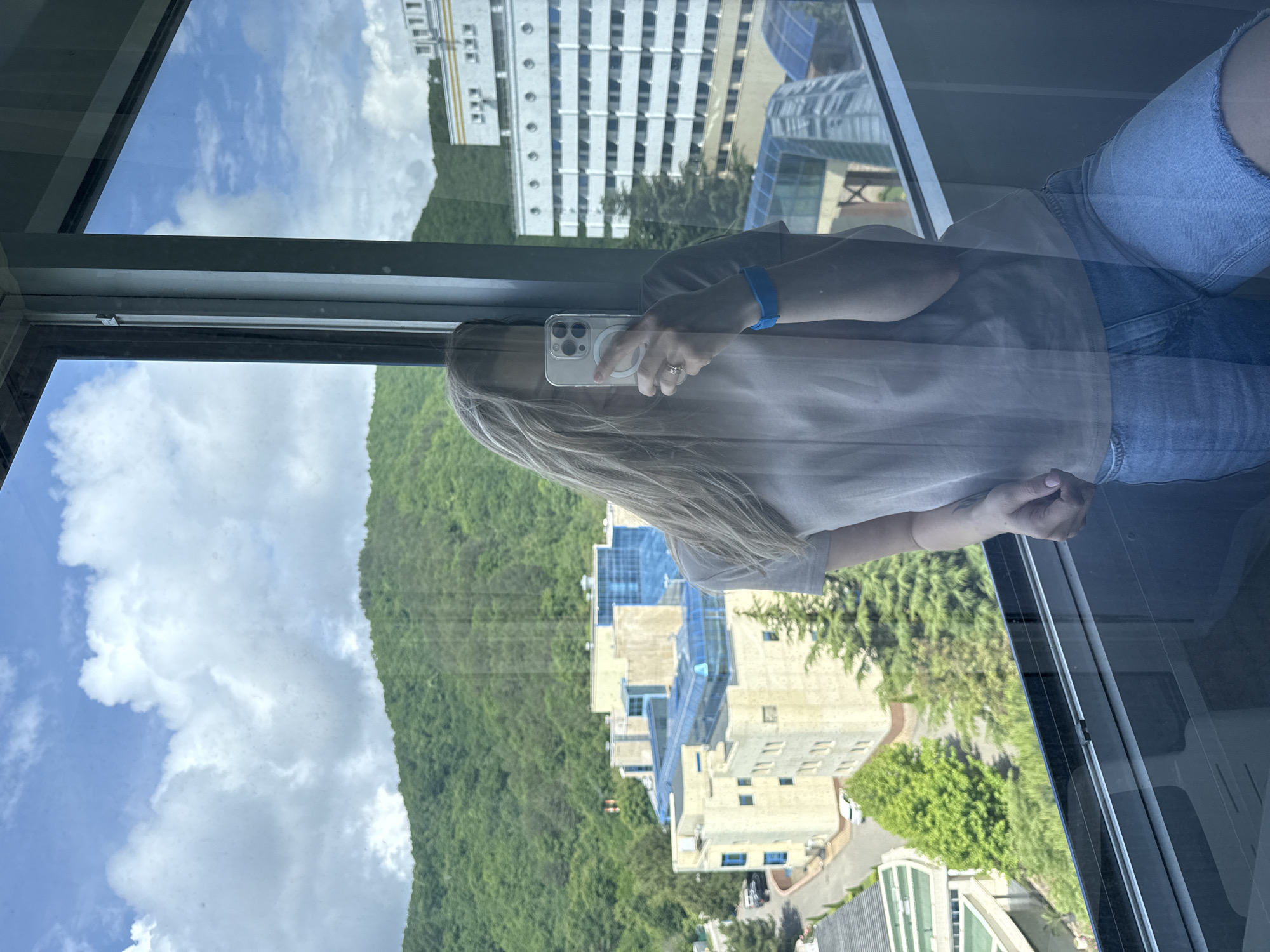 A woman with blonde hair is taking a selfie in front of a large window. She is wearing a light-colored t-shirt and denim shorts, and a blue wristband. Her face is mostly obscured by her phone and hair. Outside the window, there is a view of green, tree-covered mountains, several multi-story buildings, and a cloudy sky. The buildings appear to be part of a resort or complex. The scene is likely located in the city of Nebug, Russia, given the presence of mountains and resort-style architecture.