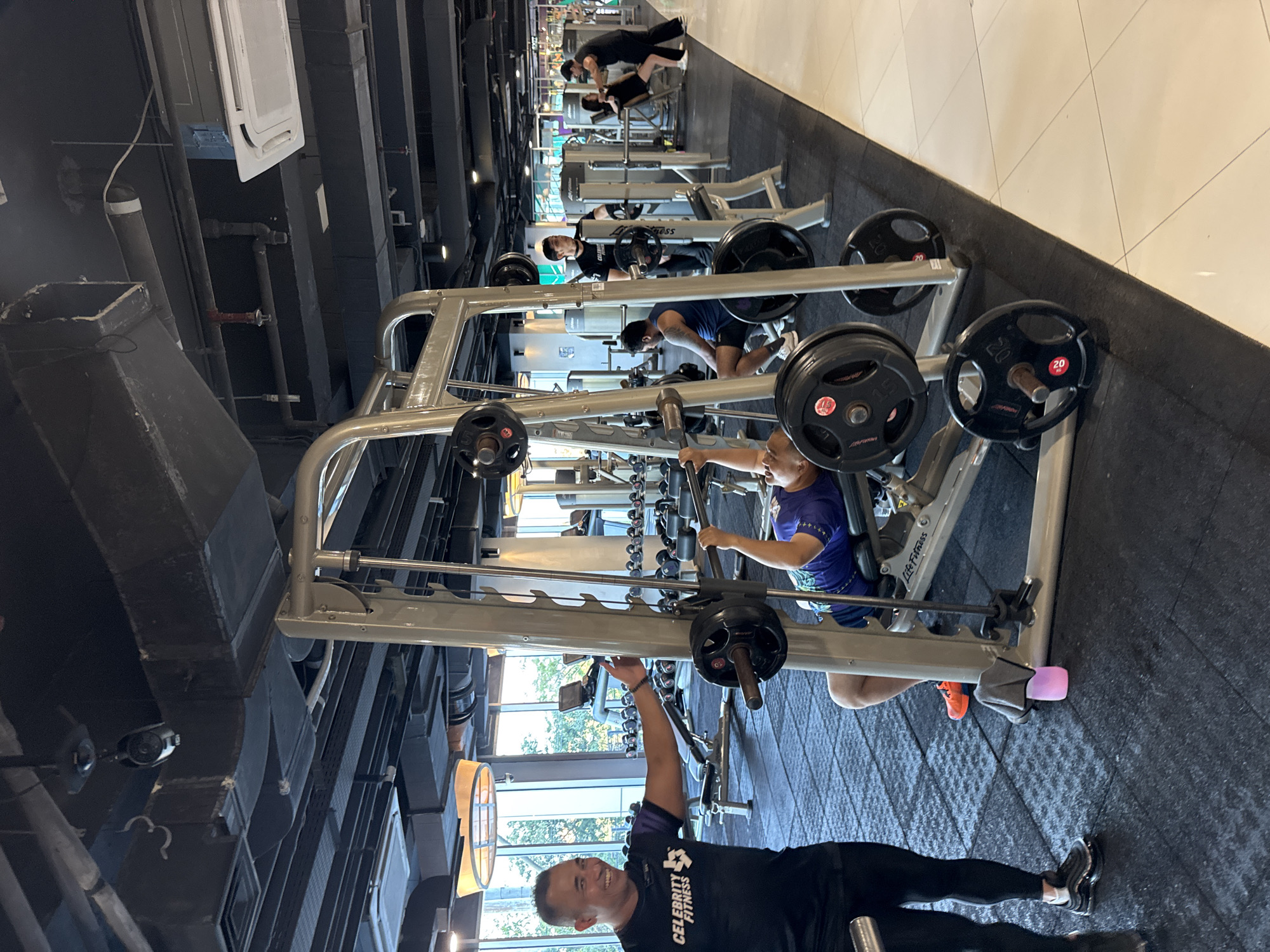 The image captures an indoor scene at Celebrity Fitness in Kuta, Indonesia. The focus is on a gym environment with multiple people engaged in various workout activities.
In the foreground, a man in a black "Celebrity Fitness" t-shirt and black leggings stands, smiling and gesturing towards a man lying on a bench press machine. This man on the bench is wearing a blue and purple patterned tank top and lifting a barbell.
Behind them, other individuals are visible. To the right, a man in a black t-shirt and shorts stands near a weight rack, observing. Further back, another man is seen spotting a woman on a different weight machine.
The gym features a variety of exercise equipment, including weight machines, benches, and racks of weight plates. The flooring is a combination of black rubber mats and white tiled surfaces. The ceiling is industrial, with exposed pipes, ventilation ducts, and lighting fixtures, including a circular pendant light visible in the background. Large windows in the background offer a glimpse of an outdoor area, possibly showing trees and more gym equipment. The overall impression is a well-equipped and active fitness center.