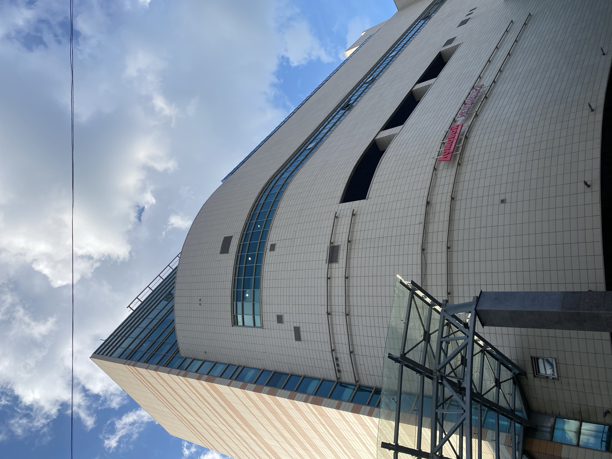 A low-angle upward shot captures a multi-story commercial building under a partly cloudy sky in Sinyavino, Russia. The building comprises distinct architectural sections; the left portion is a tall, rectangular structure featuring a facade of alternating light beige and reddish-brown horizontal bands, interspersed with vertical strips of blue-tinted reflective glass windows. The larger, central, and right sections of the building are curved and clad in light-colored square tiles, displaying multiple horizontal bands of blue-tinted windows and several dark, recessed, rectangular openings. A red sign with Cyrillic text, reading "АСНОЛБОЛ ТОРГОВЫЙ ДОМ" and "УНИВЕРСАЛ", is affixed to the tiled facade on the right side. In the lower-left foreground, a modern glass and steel canopy structure, supported by a dark, rectangular column, extends outwards, likely covering an entrance. The sky is predominantly blue with scattered white cumulus and stratocumulus clouds. A thin, dark cable or wire is visible horizontally stretched across the upper part of the frame against the sky. No individuals or active elements are present.