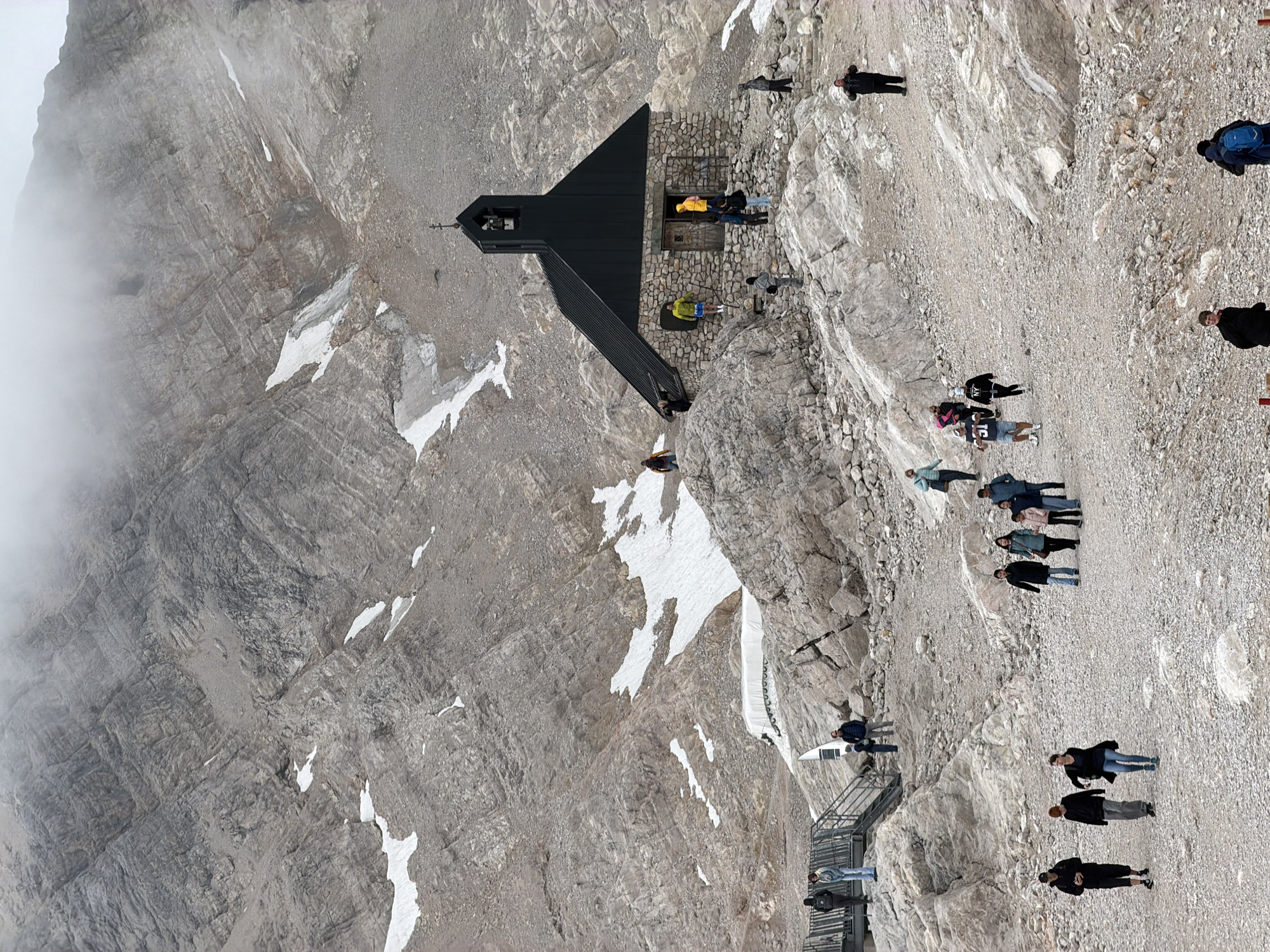 This image depicts the exterior of the small chapel, known as the "Kapelle auf dem Zugspitzplatt," located at the Zugspitzplatt plateau on Zugspitze mountain, near Garmisch-Partenkirchen, Germany. The chapel is constructed of stone with a dark, pitched roof and a small bell tower. Several tourists are visible in the scene, some walking on the rocky, gravelly terrain, others gathered near the chapel or on pathways. The mountain landscape is rugged and rocky, with patches of snow scattered across the slopes, and the sky is overcast with mist obscuring the higher peaks. The overall impression is of a high-altitude tourist destination with a prominent religious structure.