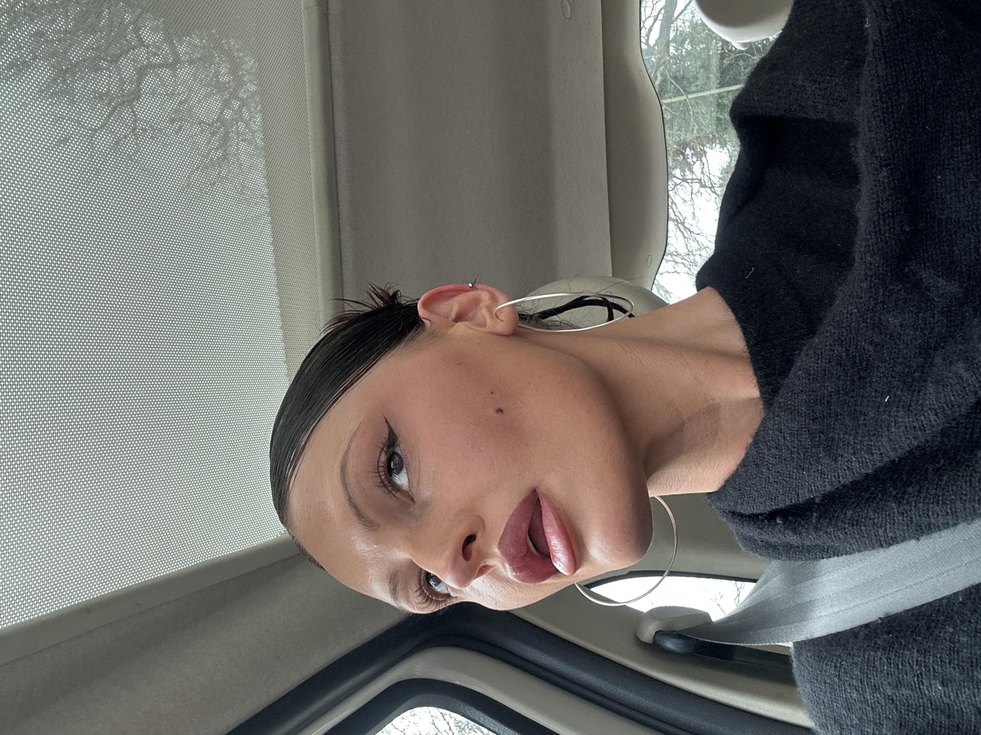A female individual is seated in the passenger compartment of a vehicle within the Wealden district, United Kingdom. She wears a black garment and a grey car seatbelt diagonally across her chest and left shoulder. Her dark hair is slicked back. Her face displays prominent makeup, including winged eyeliner, defined eyebrows, and glossy, slightly parted lips. A small dark mole is visible on her left cheek. She wears a large silver hoop earring and a small upper cartilage piercing in her left ear, looking upwards and to the right. The vehicle interior features a light-colored headliner and a panoramic sunroof with a perforated screen. Through the windows, bare tree branches under an overcast sky are visible, indicating an outdoor, non-urban environment.