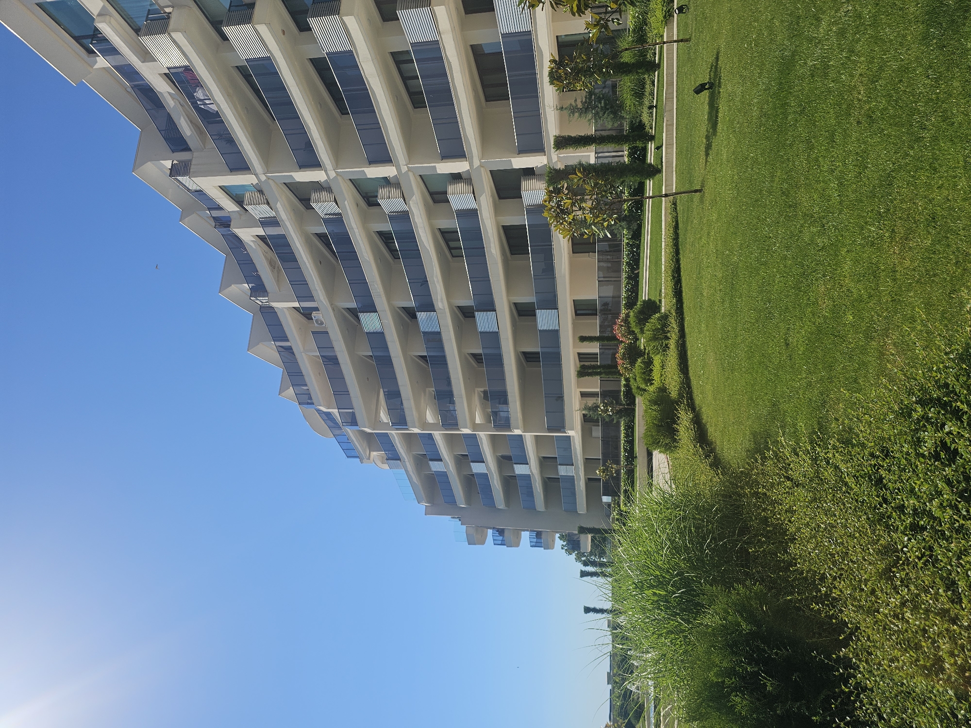 An exterior view of a modern, curved apartment building with white facades and balconies with glass railings is captured under a clear blue sky. The building has multiple stories and a stepped roofline. In the foreground, a well-maintained lawn with manicured bushes and some ornamental trees is visible. Tall, reedy green grass grows along the left side of the frame. The scene appears to be a residential or resort area.