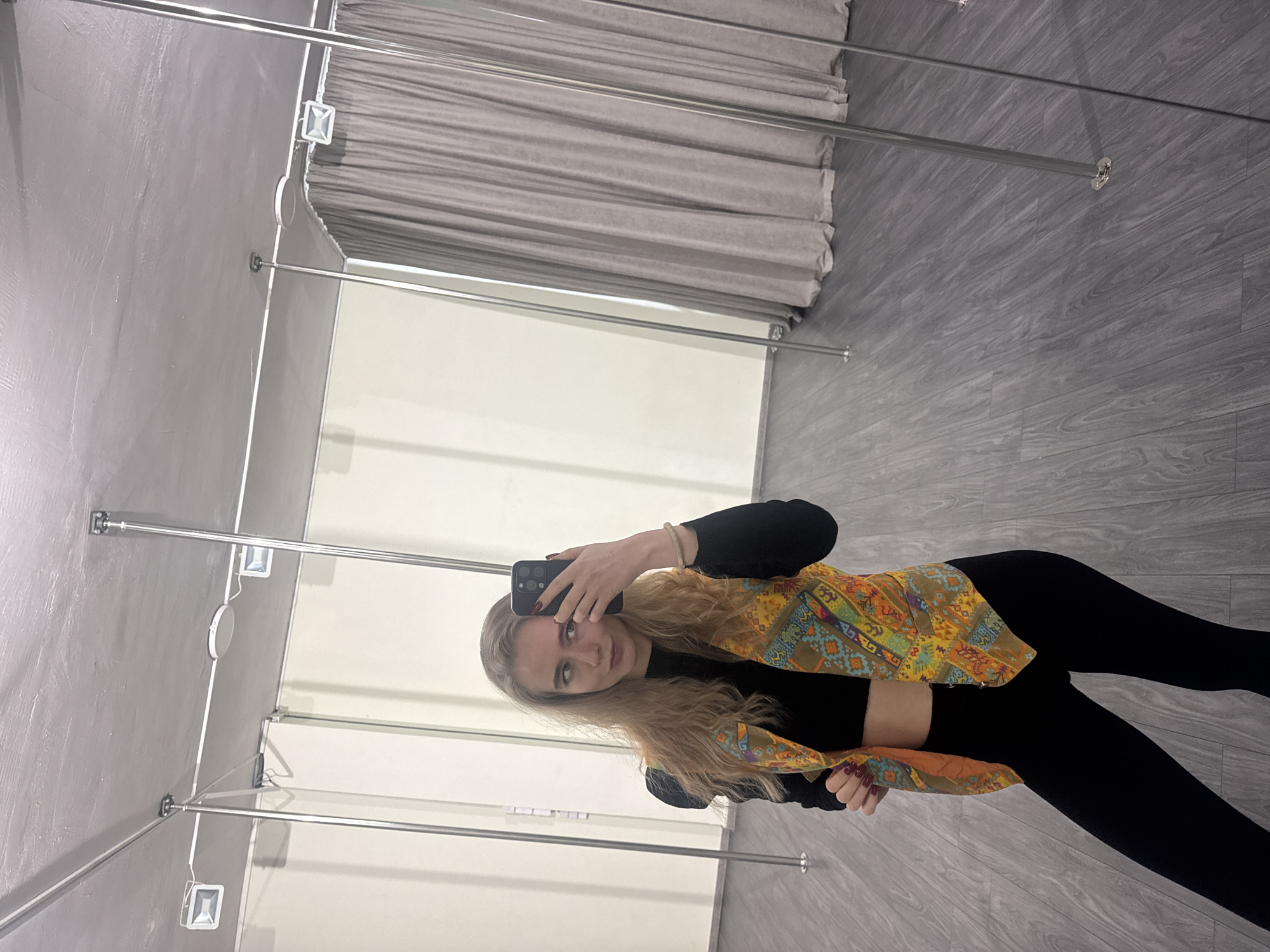 A young adult female is depicted taking a mirror selfie in what appears to be a pole fitness or dance studio located in the city of Unknown, Unknown. The subject, with long wavy blonde hair, is dressed in a black long-sleeved crop top, black leggings, and an open, multi-colored patterned vest featuring geometric and floral designs. She holds a dark-colored smartphone in her right hand, positioned to capture her reflection, while her left hand is near the bottom of her vest. Her fingernails are painted dark red, and a light-colored beaded bracelet is on her right wrist. The room features grey wood-grain laminate flooring and a grey, textured ceiling from which multiple polished chrome vertical poles extend. The walls are predominantly light beige, with a large section covered by light grey pleated curtains on the right side of the frame. Two rectangular white LED floodlights and one circular light are mounted on the ceiling. The entire scene is reflected in a large mirror.