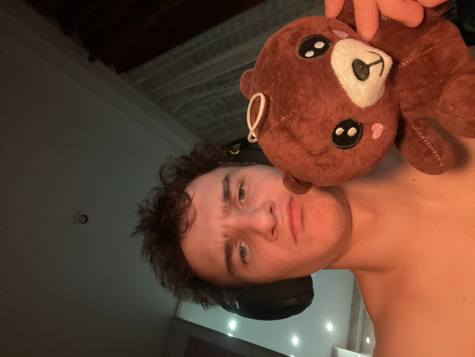 A light-skinned person with curly brown hair, wearing black headphones, holds a stuffed brown bear with embroidered features near their head. The individual has a neutral expression. The background shows a dimly lit room with a slatted ceiling and a dark wall on the left. The person appears shirtless or wearing a very low-cut garment.