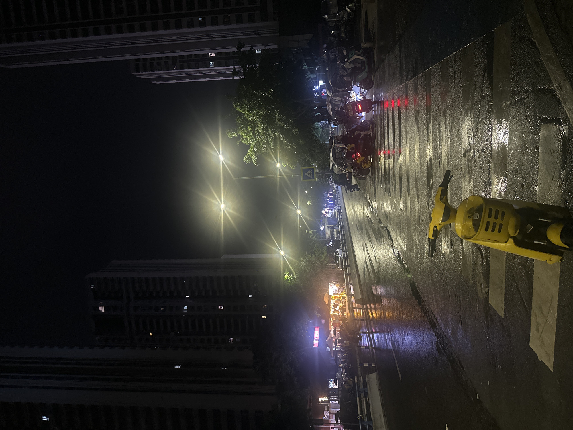 This image depicts a nighttime urban street scene in Chongqing High-tech Zone, China, following recent rainfall, as indicated by the wet, reflective asphalt. Tall, multi-story buildings, characteristic of modern Chinese cities, line both sides of the street, with a number of illuminated windows visible against the dark sky.
In the foreground, a bright yellow shared electric scooter, facing away from the viewer, is positioned on a pedestrian crosswalk. The street itself is an asphalt road with visible white pedestrian crossing stripes and a wet surface that strongly reflects the artificial lighting.
Mid-ground to background on the left, a cluster of brightly lit market stalls or food vendors operates under various awnings, creating a focal point of activity. Several individuals are present around these stalls, and their reflections are visible on the wet pavement. A blue guardrail separates the pedestrian area from the main thoroughfare.
On the right side of the street and extending into the mid-ground, numerous parked and stationary electric scooters, motorcycles, and three-wheeled enclosed motor-rickshaws (sanlunche) are present. Several of these vehicles have visible red taillights illuminated. People are seen riding or standing near some of these vehicles.
Multiple streetlights, emitting distinct starburst patterns, illuminate the scene, casting strong reflections on the wet road surface. Trees with green foliage are visible along the sides of the street, some partially illuminated by the artificial light. A pedestrian crossing sign is visible near a tree in the mid-ground. The overall impression is a bustling urban environment at night.
