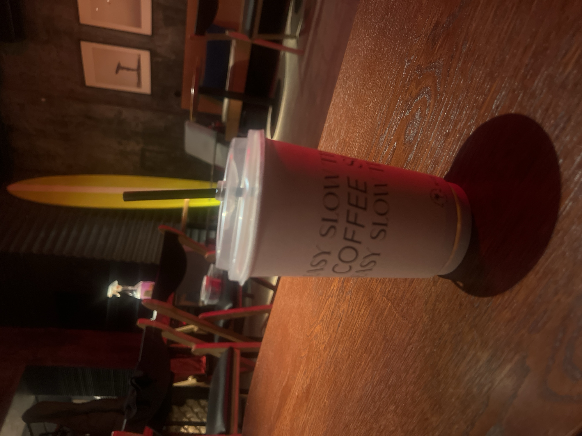 A white and red paper coffee cup from Easy Slow Coffee in Moscow, Russia, sits on a wooden table. The cup has a clear plastic lid and a black straw. The table is brown and has a visible wood grain. The background includes chairs and a yellow surfboard. The room is dimly lit.