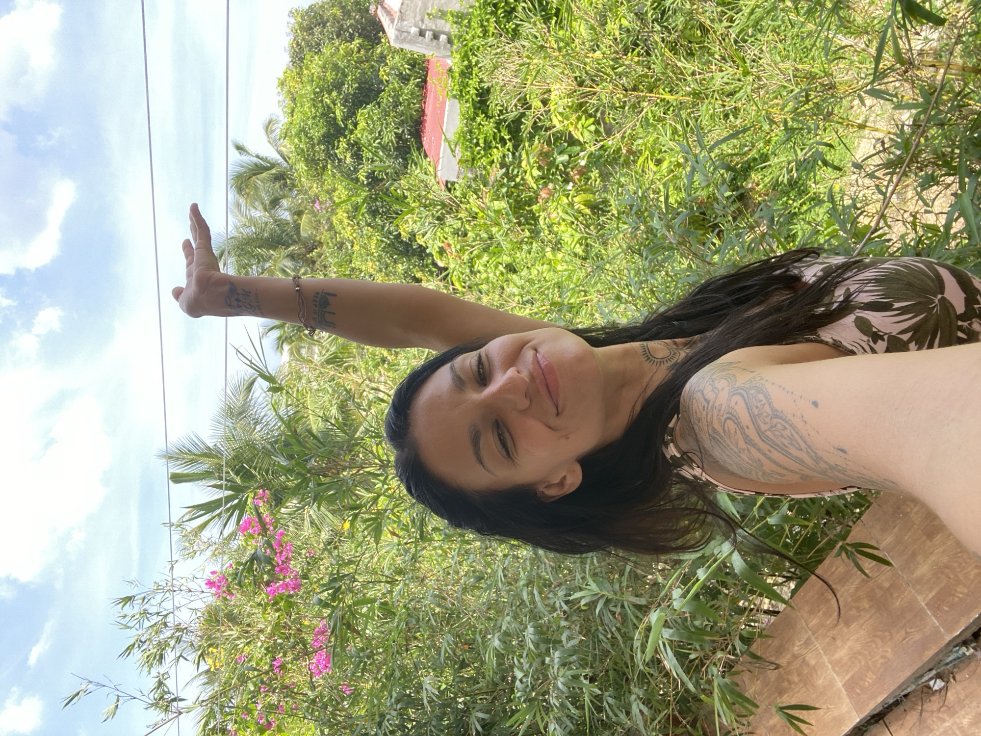 An outdoor, medium shot captures a woman with dark hair, smiling and holding her right arm up with an open palm. She is positioned in the foreground, with lush green foliage and bamboo plants surrounding her. In the background, palm trees and a red-roofed building are visible under a blue sky with scattered clouds. The woman has tattoos on her left arm and chest. This scene is likely in Mũi Né, Vietnam, suggested by the presence of tropical vegetation and architecture typical of the region.