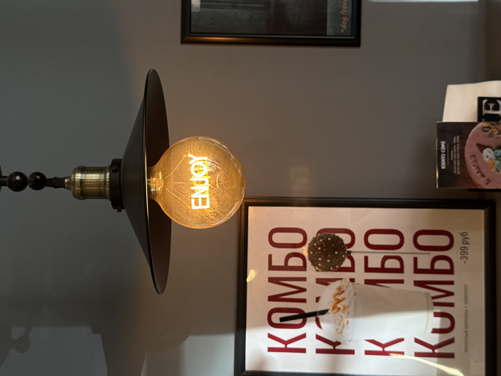This vertically oriented indoor photograph captures a cozy and inviting atmosphere, likely within a cafe or restaurant in Krasnoyarsk, Russia. The central subject is an illuminated, decorative Edison-style light bulb, whose glowing filament clearly spells out the word "ENJOY." This bulb is nestled within a stylish black metal lampshade, suspended from a dark, articulated fixture against a neutral grey wall.
To the right, a framed advertisement hangs, featuring a tall, frothy drink (possibly a milkshake) with caramel drizzle and whipped cream, alongside a chocolate cake pop on a stick. The prominent Cyrillic text on the poster reads "КОМБО" (Combo), repeated as a background pattern. Smaller text identifies the offering as "молочный коктейль + кейкпопс" (milkshake + cake pop) and lists the price as "-399 руб" (399 rubles).
Above and to the right of the framed advertisement, a stack of papers or menus is partially visible. Text on these items includes "IBENTO CAKE" and "*dog fren" (likely indicating a dog-friendly establishment). The warm, artificial light from the bulb creates a soft glow, suggesting the shot was taken indoors.