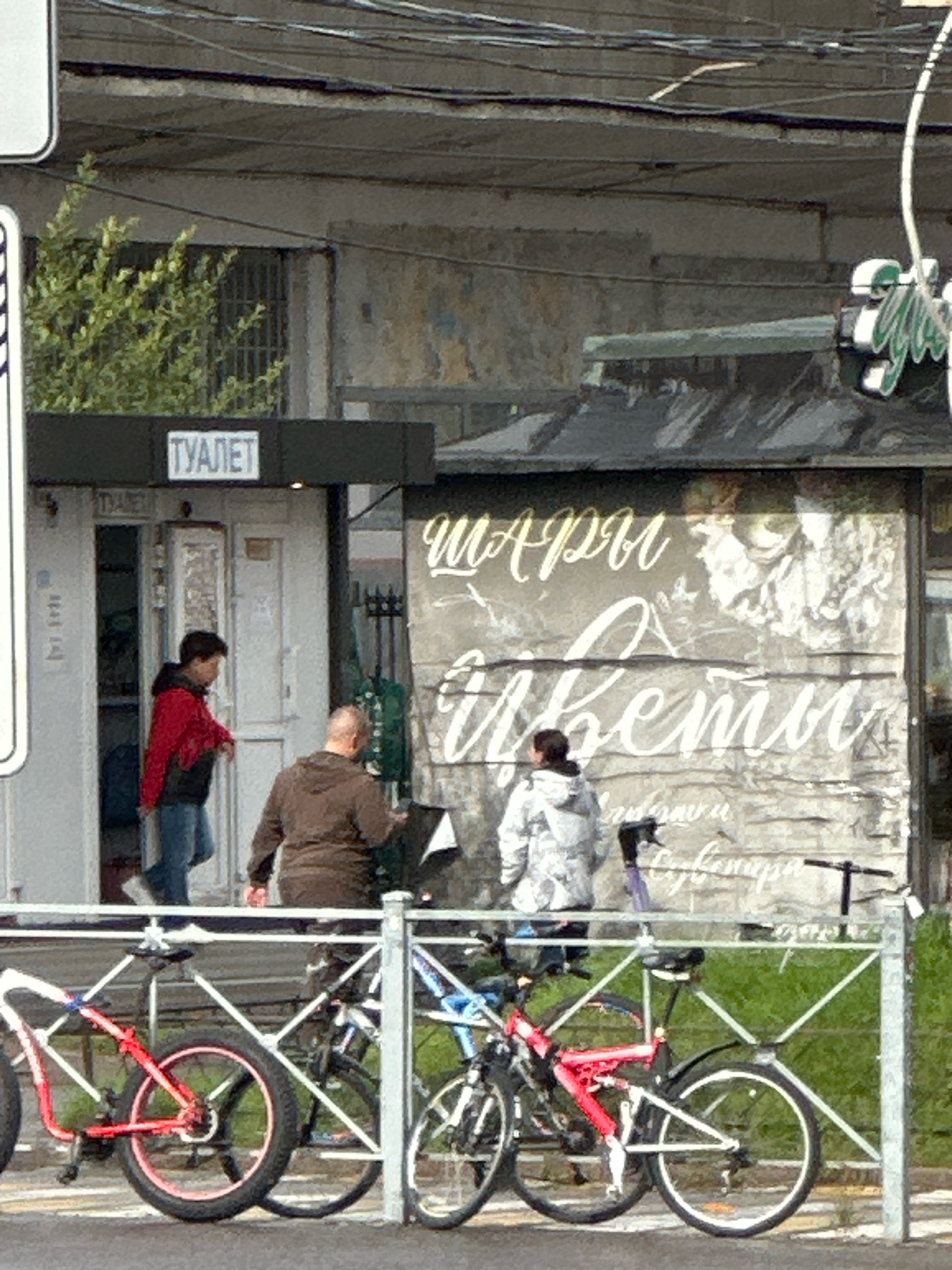 In Murino, Russia, on a bright, overcast day, a scene unfolds outside a small building with a sign reading "Туалет" (Toilet). Two men are present. One man, wearing a red jacket and blue jeans, is exiting the building. The other man, in a brown hooded jacket, is walking away from the building towards the right of the frame. To the right of the building, a wall is adorned with large, faded white lettering that appears to read "Мари Цветы" (Mary Flowers).
In the foreground, a white metal fence separates the sidewalk from a grassy area. Parked behind the fence are several bicycles. A red bicycle with thick tires and pink rims is prominent on the left, while other bicycles, including a red and black one and a blue and white one, are visible further to the right. One person, wearing a grey and white patterned hooded jacket, is seen near the bicycles, potentially interacting with them or the environment. The overall scene suggests a public space with everyday activities taking place.