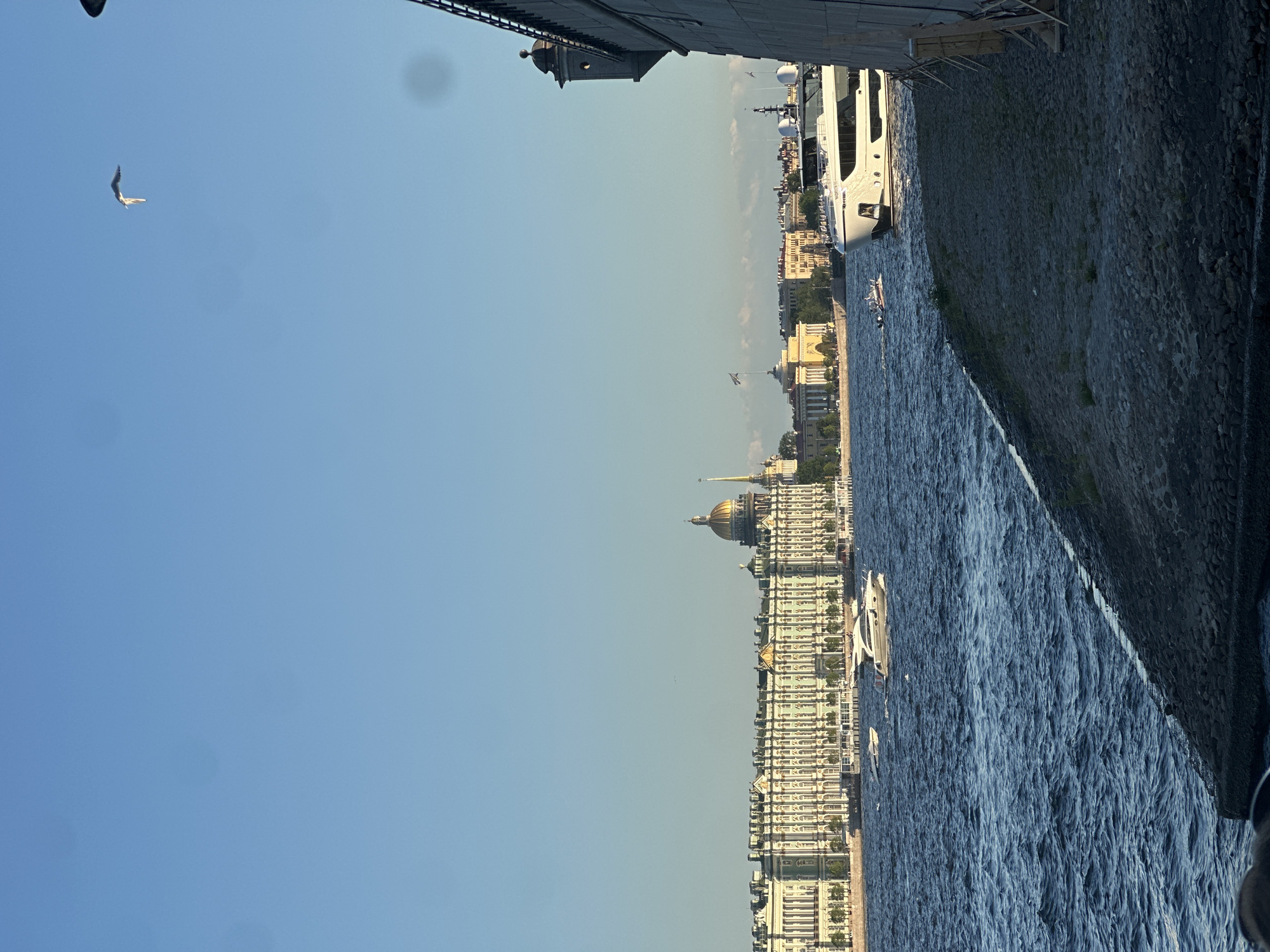 This image depicts a panoramic view of the Neva River in Saint Petersburg, Russia, featuring prominent architectural landmarks and maritime activity.
In the background, dominating the left side of the frame, is the iconic Winter Palace, home to the Hermitage Museum. It is a long, multi-story building characterized by its pale green and white facade, numerous arched windows, and decorative classical elements. Behind it, a distinctive golden dome (likely Saint Isaac's Cathedral) and a very tall, slender golden spire (likely the Admiralty building) are visible against the clear sky. Further to the right, across the river, several yellow neoclassical buildings with flags can be seen, establishing the cityscape along the embankment.
The midground is occupied by the dark blue, choppy waters of the Neva River, reflecting the sunlight in various ripples. Several boats are present: a large, modern white motor yacht, partially obscured by the foreground wall, is prominently featured on the right. Its name "WASHINGTON" is partially visible on its bow. Another similar white motor yacht is moving further left, creating a slight wake. A smaller, dark-hulled speed boat with several indistinct figures aboard is also in motion on the water.
The foreground consists of a dark, textured stone embankment or wall, composed of irregular grey stones. On the upper right, a dark, small turretted structure, possibly part of a fortress or bridge support, protrudes from the wall. Towards the bottom right, several wooden planks or supports are propped against the embankment.
The sky above is a clear, bright blue with no visible clouds. A single white bird, likely a seagull, is captured in flight in the upper right quadrant, its underside illuminated by the sun. Several soft, circular dark smudges or lens flares are visible in the upper and middle portions of the sky. The overall lighting suggests late afternoon or early evening, with warm sunlight illuminating the buildings and the bird.