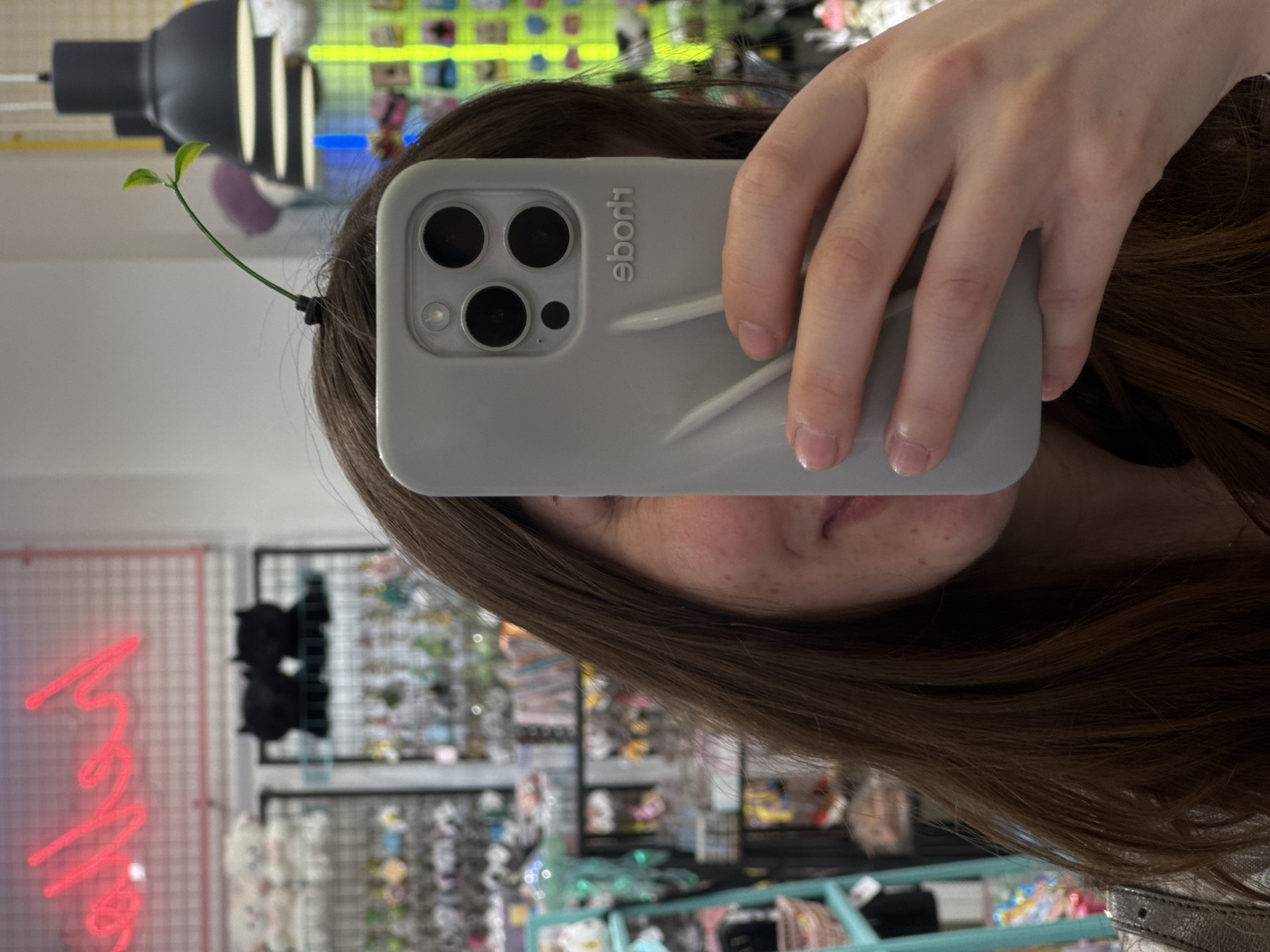 A person takes a selfie with a grey iPhone. A small green plant sprouts from the person's hair. In the background are shelves of merchandise and a pink neon sign. The location is not identifiable as Moscow, Russia from the visual information provided.