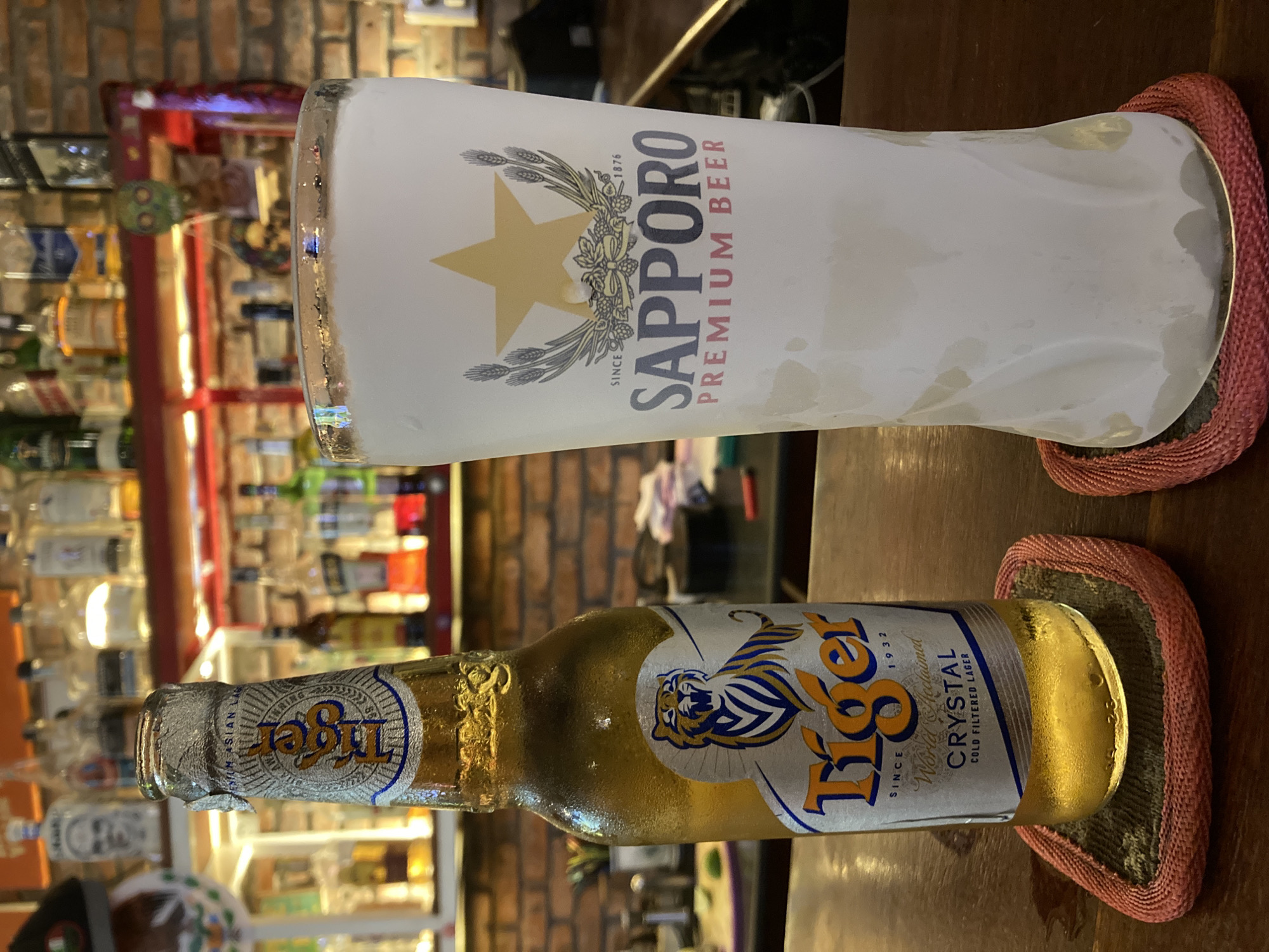 The image shows a Sapporo Premium Beer glass and a bottle of Tiger Crystal beer. The glass is tall and frosted, displaying the Sapporo logo with the gold star and the phrase "SINCE 1876 SAPPORO PREMIUM BEER." The Tiger Crystal bottle, with a gold liquid inside, has its label clearly visible with the tiger logo and the words "SINCE 1932 World Acclaimed CRYSTAL COLD FILTERED LAGER." Both the glass and the bottle are resting on red coasters, placed on a wooden surface. In the blurred background, there is a partially visible brick wall, a shelf stocked with numerous bottles, and other bar-like fixtures. It is probable that the location is in Phu Thuy Ward, Vietnam.