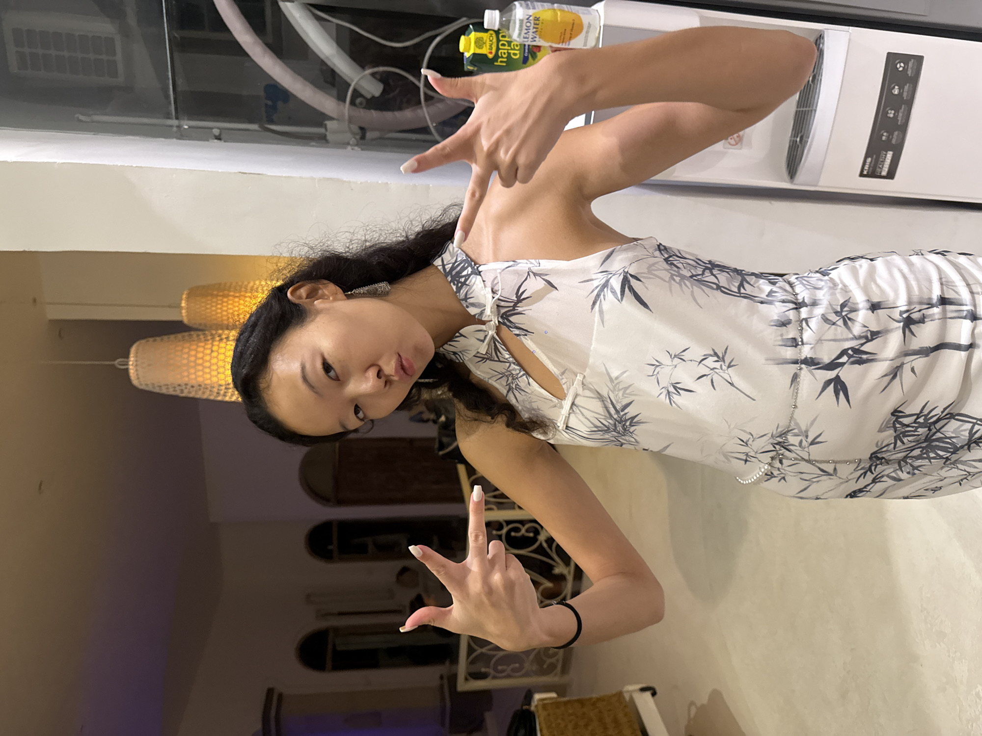 In North Kuta, Indonesia, a young woman with dark hair poses for a photo. She is wearing a white dress with a bamboo print and has her nails painted white. She is making a hand gesture with both hands, holding up two fingers on each hand. The background is dimly lit and shows a room with decorative light fixtures and a water dispenser.