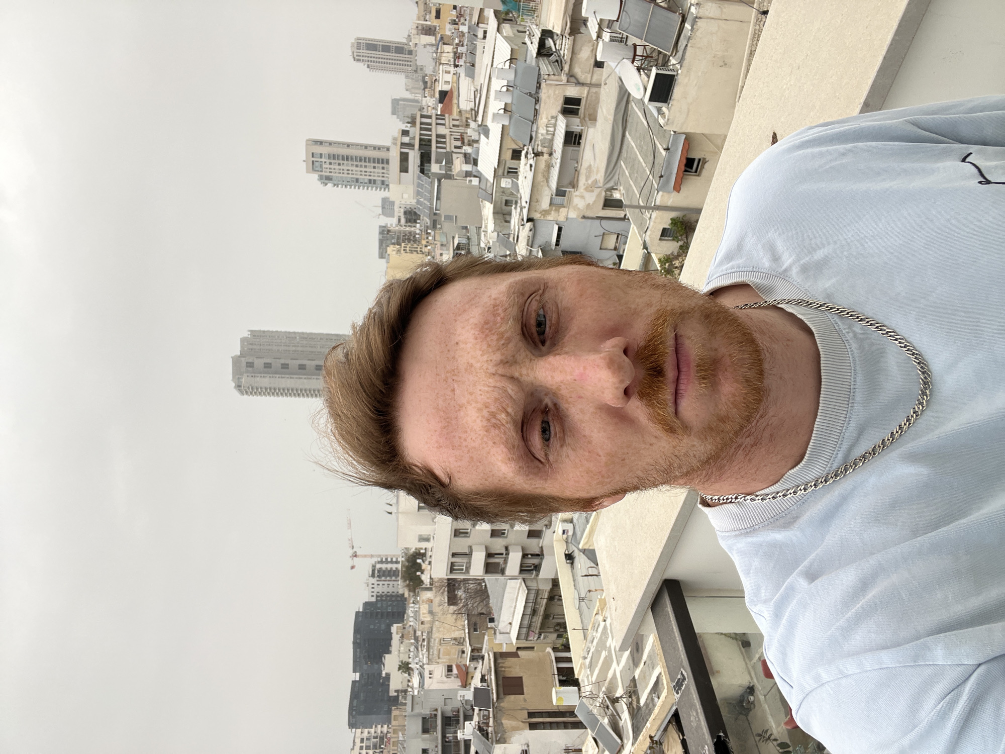 A close-up selfie shot, oriented vertically, shows a fair-skinned man with red hair and freckles on a rooftop in Tel-Aviv, Israel. He wears a light blue t-shirt and a silver chain necklace. Behind the man, the scene is dominated by a densely packed cityscape of Tel-Aviv. A mix of older, low-rise buildings with white or light-colored facades and solar panels are juxtaposed with taller, modern buildings. A crane is visible in the distance, indicating ongoing construction. The sky is overcast and grey, contributing to the muted color palette of the image. The man's face is the primary focus, with his expression appearing neutral.