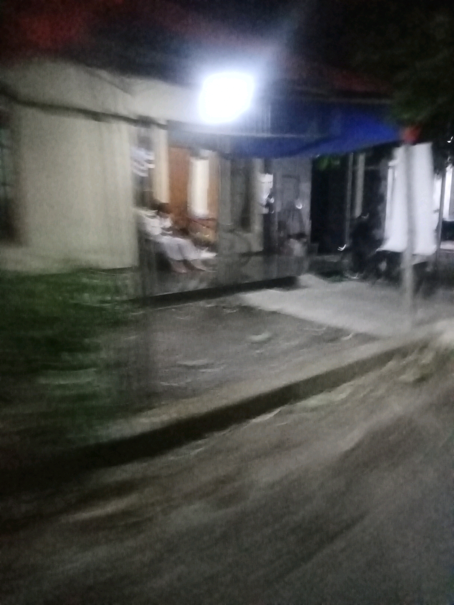 An outdoor scene at night in Sumber Mulyo, Indonesia, shows a dimly lit area with a building on the left and a covered outdoor space on the right. A bright light is visible above the covered area, illuminating a blue tarp. Several figures are present within the covered space, appearing to be seated or lying down. The ground in the foreground is a paved surface, possibly a road or walkway, with some grassy area to the left of the building. The overall image is blurry, suggesting movement or low light conditions.