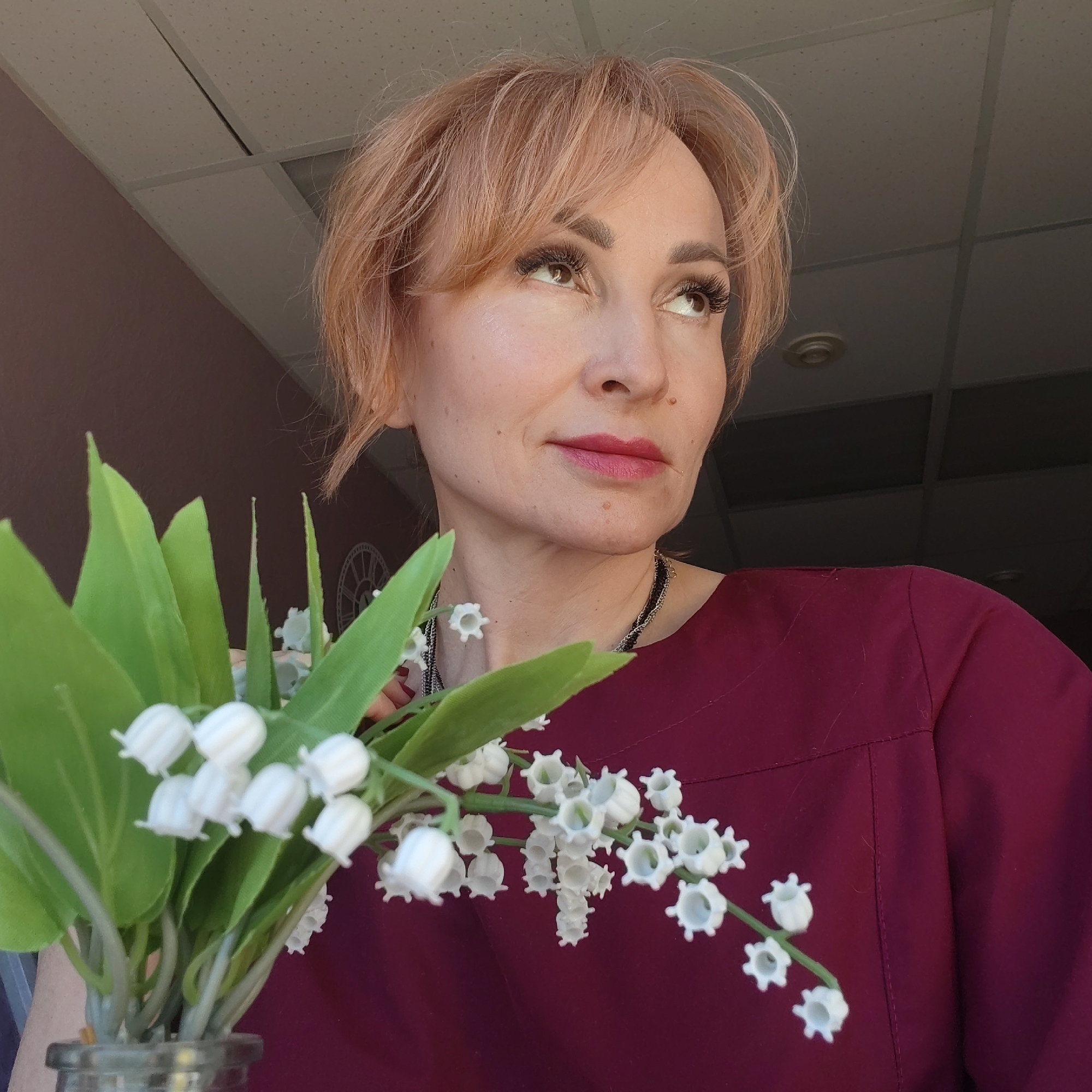 A woman with short, light reddish-blonde hair is holding a bouquet of lilies of the valley. She is wearing a dark red-purple top. The woman is looking up and to the right. The background includes a ceiling with tiles and a wall that appears to be pinkish. The image appears to be taken indoors. There is no information available within the image or prompt to verify the location in the city of Chaikovsky, Russia.