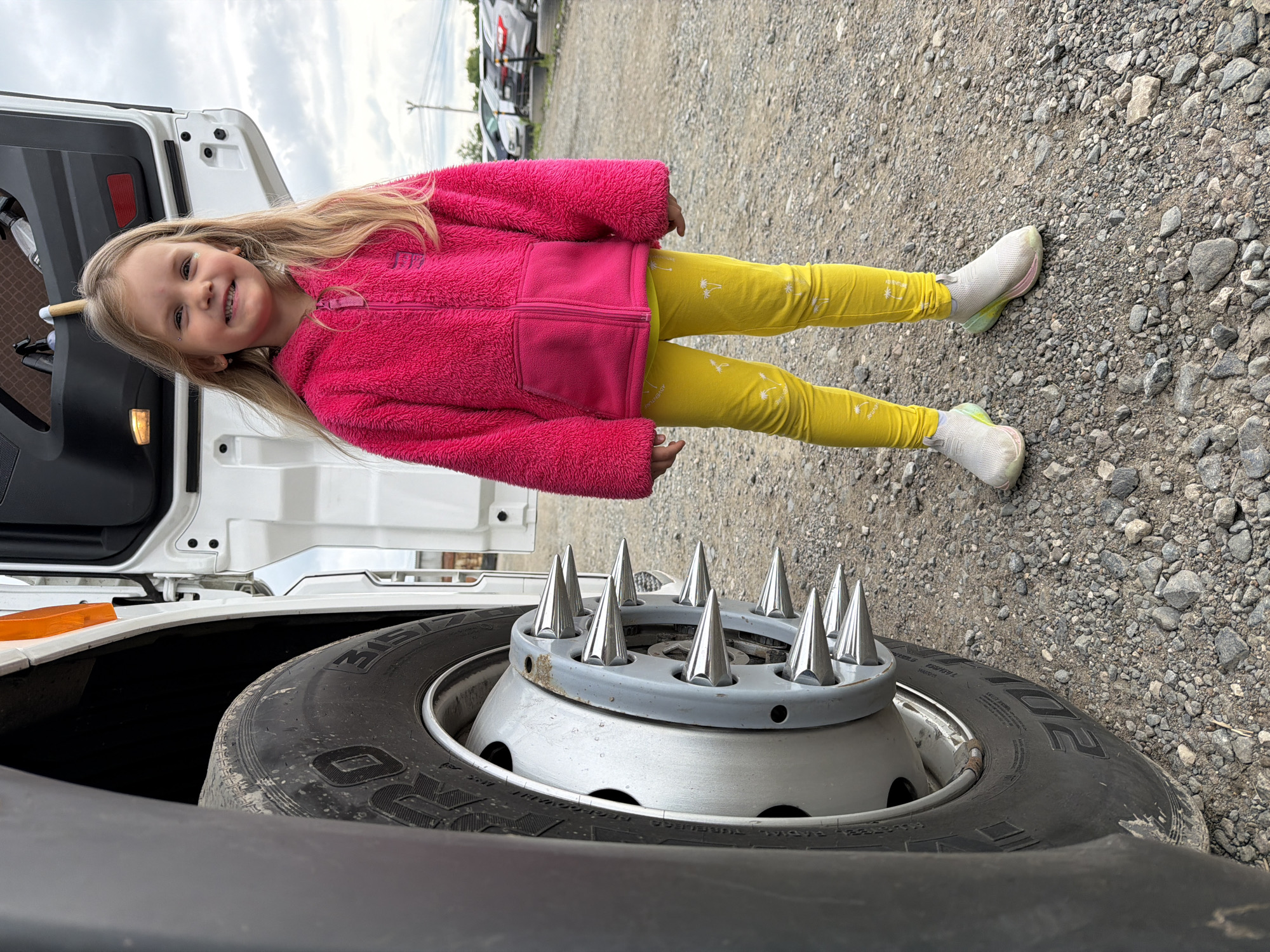 The image captures a young girl standing next to a large truck tire in an outdoor setting in Kamyennoye, Russia. The girl, with long blonde hair, is wearing a bright pink fuzzy zip-up jacket and vibrant yellow pants adorned with small white palm tree patterns and the faint text "ADVENTURE." She is smiling broadly at the camera, conveying a happy and cheerful mood. Her shoes are white athletic sneakers with light green and pink accents on the soles.
A prominent feature in the foreground is a large black truck tire, fitted with a silver hubcap that is decorated with multiple chrome spikes. Part of a white truck structure, possibly the cab or a storage area, is visible behind the girl. The ground is a rough mix of gravel and small stones, suggesting a parking lot, truck stop, or unpaved industrial area.
The sky is overcast, indicating a cloudy day. In the background, several other vehicles, possibly trucks or vans, are parked. Visible text on the truck tire includes "315/80 R22.5," indicating its size specification.