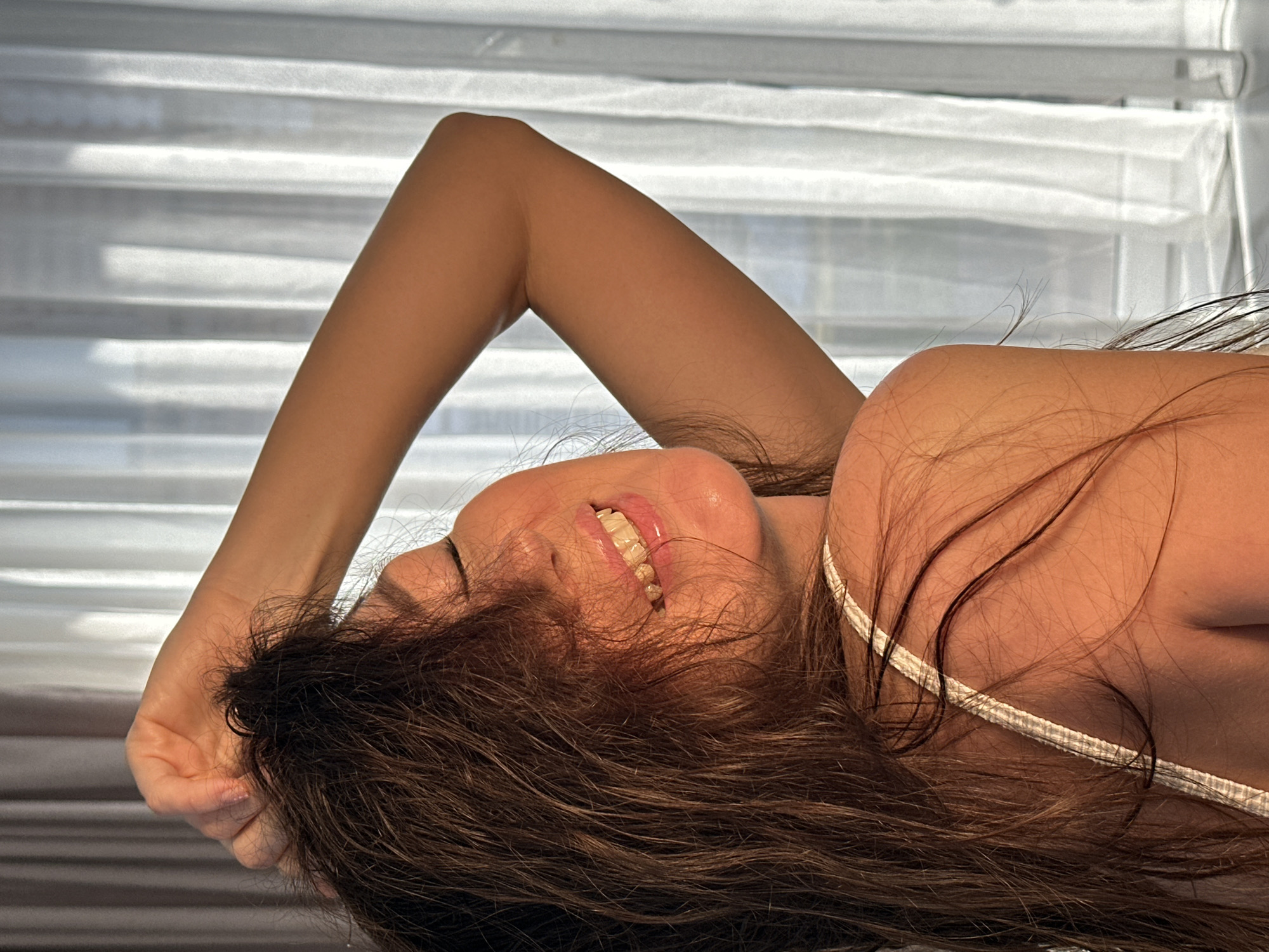 A close-up, rotated image features a person, likely a woman, lying down with her head tilted back. She is captured in a candid moment, smiling or laughing with her eyes closed and mouth open, revealing her teeth. Her long, dark brown hair frames her face and spreads around her shoulders. One arm is raised and bent, with her hand touching her head or hair, while her other shoulder and a thin, light-colored strap of a top or undergarment are visible in the foreground.
The background consists of brightly lit horizontal blinds or curtains, indicating an indoor setting with natural daylight. The lighting highlights the person's warm skin tone and the texture of her hair. The emotion conveyed is one of happiness, joy, or relaxed amusement. No text is visible in the image. The scene is set in Chaikovsky, Russia.