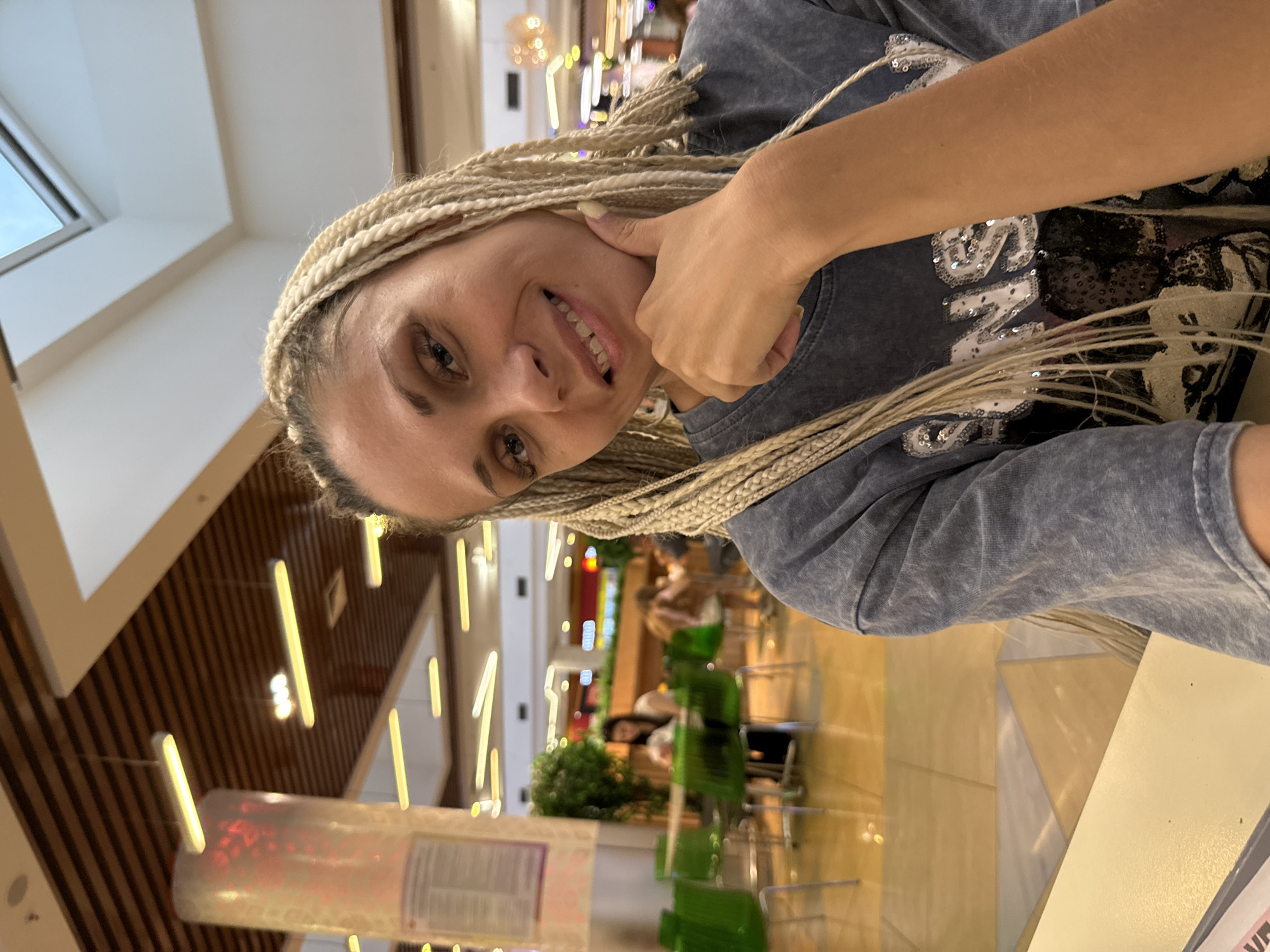 The image shows a young woman with blonde braided hair smiling and resting her chin on her hand. She is wearing a dark gray t-shirt with a sequined design. She is seated at a table, and the background suggests an indoor public space, possibly a food court or shopping mall. There are other people and tables visible in the blurred background, as well as bright overhead lighting. The setting appears to be a modern, well-lit interior. The image does not contain any visual information that would indicate the location is in Arkhangelsk, Russia.