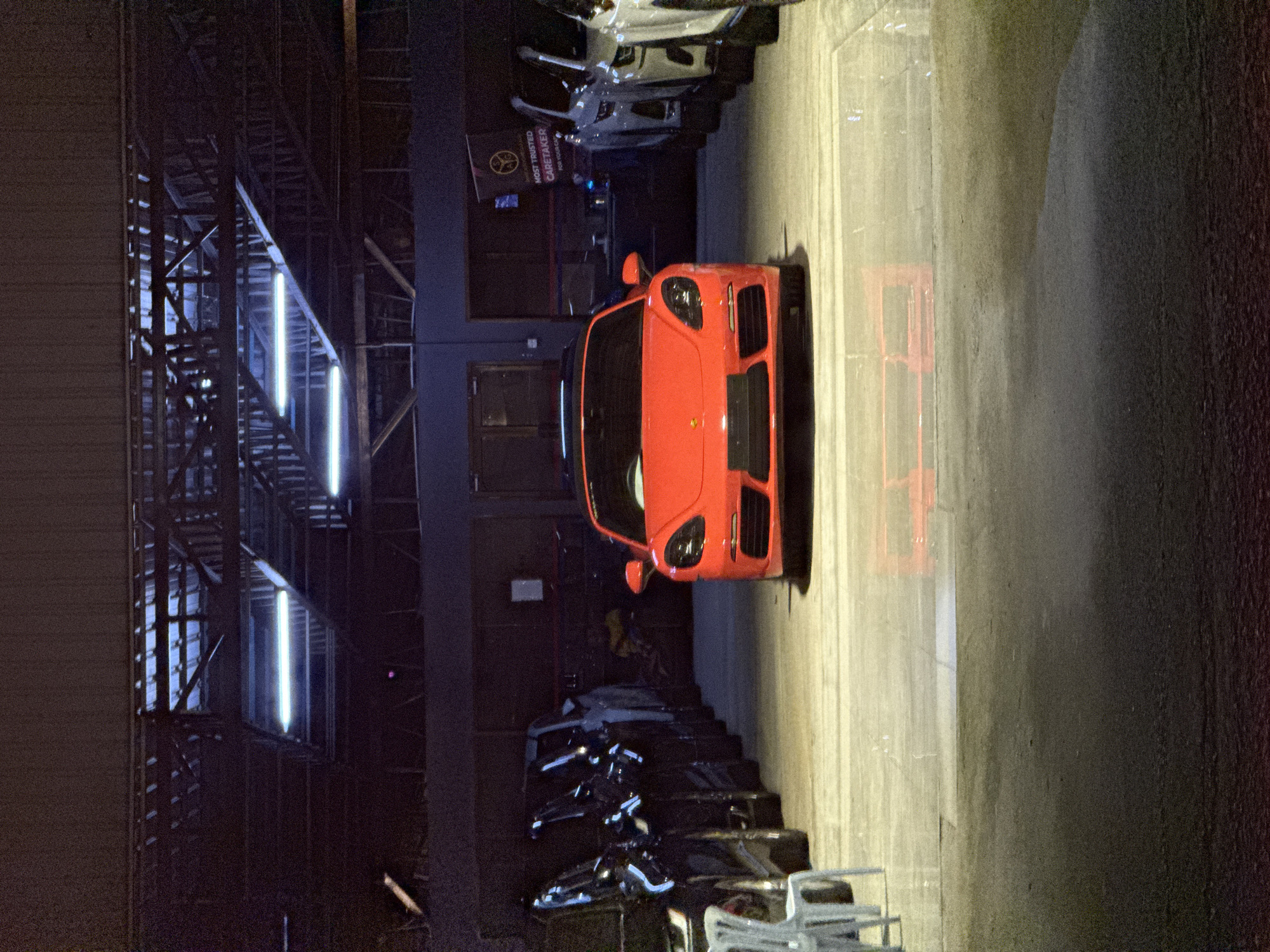 The photograph depicts a warehouse or large garage-like space, possibly in Kuala Lumpur, Malaysia, though specific landmarks are absent to confirm. The foreground features a bright orange sports car parked on a concrete floor, its front end facing the viewer. Behind the car are several other vehicles, possibly motorcycles and cars, parked in a row. The ceiling of the warehouse is high and exposed, with metal beams and visible fluorescent lighting. A sign is partially visible on the wall behind the sports car. The lighting is a mix of bright artificial light from the ceiling fixtures and darker areas in the corners and edges of the space. The floor appears clean and well-maintained.