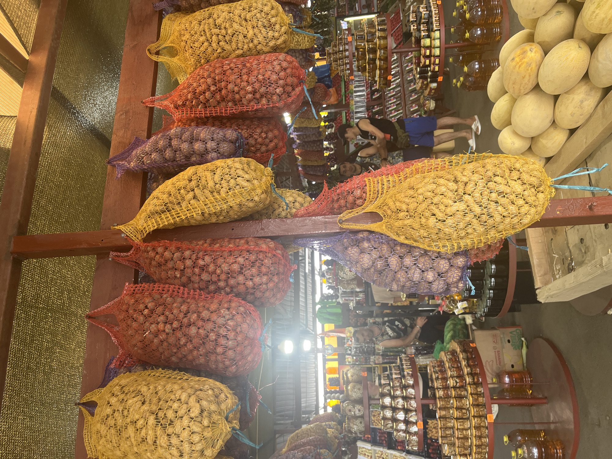 This is a photograph taken outdoors at a market stall in Plastunovskaya, Russia. The stall is covered by a yellow-green mesh tarp, providing shade. The foreground is dominated by several large mesh bags filled with produce. From left to right, a large yellow mesh bag contains what appear to be potatoes or onions. Next to it, a red mesh bag holds a similar type of produce, followed by another red mesh bag. A yellow mesh bag filled with the same produce is hanging in the center. To the right, a purple mesh bag is partially visible, followed by another red mesh bag, and then a yellow mesh bag.
In the background, a market stall displays various goods. On shelves, numerous jars of preserved food, likely pickles or jams, are neatly arranged. Several large plastic bottles filled with amber-colored liquid, possibly oil or juice, are also visible. Two men are standing in the middle ground, appearing to be interacting with each other. One man, wearing a black tank top and blue shorts, is looking towards the camera. The other man, in a patterned sleeveless shirt and dark shorts, is facing away from the camera. Further in the background, more market stalls and produce can be seen. A pile of melons is visible in the lower right corner of the image.