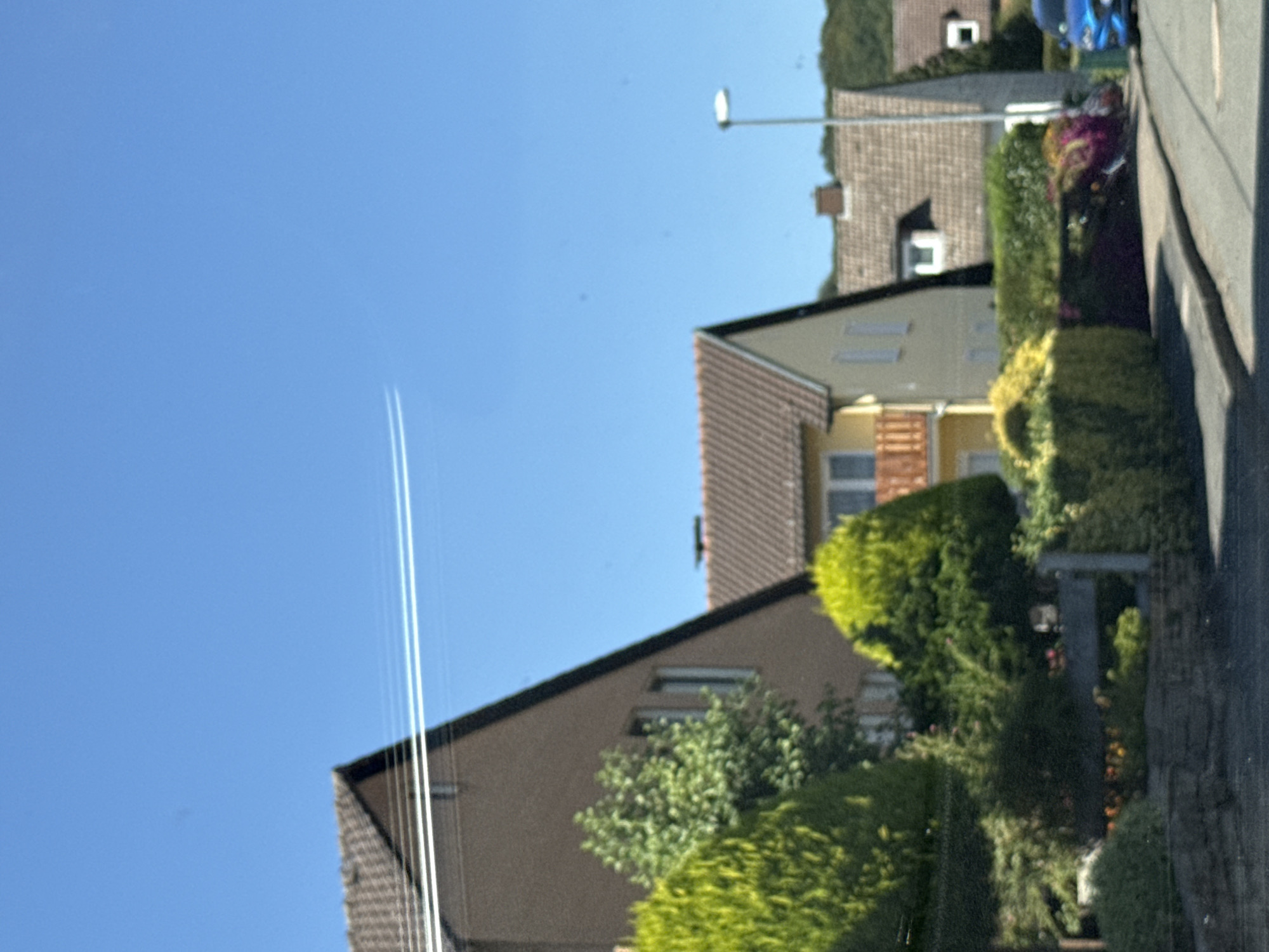 The image shows a suburban street in Bad Sachsa, Germany, under a clear blue sky. Several houses with tiled roofs are visible, with various types of landscaping, including manicured bushes and trees, in front of them. A street lamp stands on the right side of the road, and a blue car is parked on the right. In the sky, two distinct white contrails from an aircraft are visible, stretching horizontally across the frame. The scene depicts a typical residential area on a bright, sunny day.