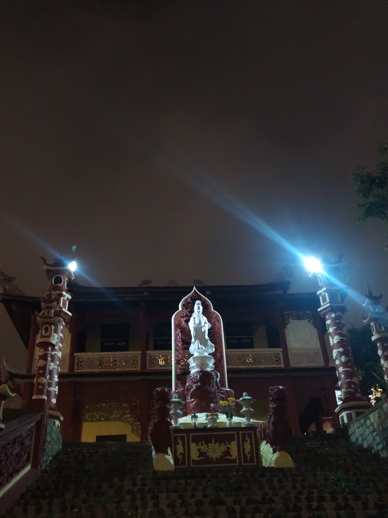 At night, the image features a reddish-brown building with a white statue centered in front. The building has a traditional Asian style, with an overhanging roof, ornate decorations, and a balcony. Flanking the statue are two red lion statues. The white statue is of a figure standing on a pedestal.
Two illuminated pillar-like structures stand to either side of the building. Light flares emanate from the top of these structures. The foreground shows stone steps leading up to the building. The sky is dark and overcast. Some greenery can be seen on the right side of the frame.