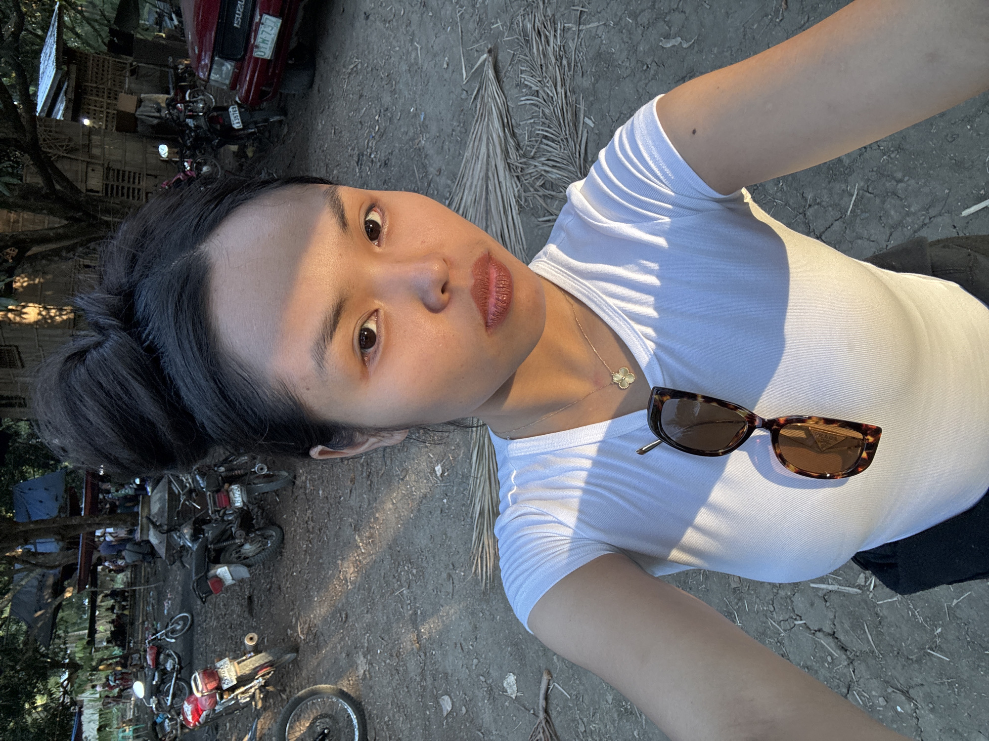 A young woman is taking a selfie outdoors in Kiblawan, Philippines. She has dark hair pulled back, wears a white top and a gold necklace, and sunglasses rest on her shirt. The background shows a rural area with motorcycles, a red pickup truck, and simple dwellings under trees. The ground is dry and cracked.