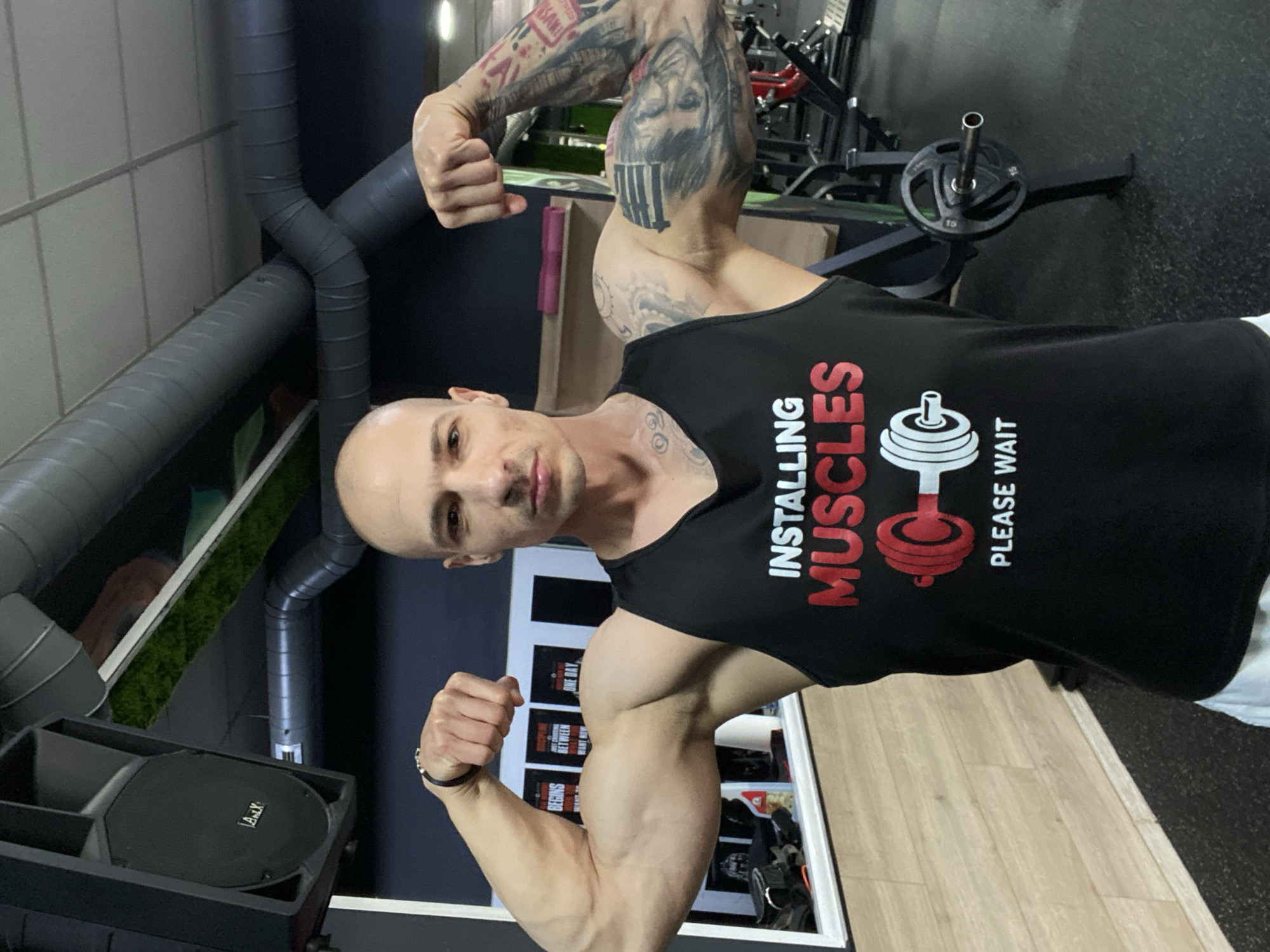 A bald man with multiple tattoos on his arms poses in a gym, flexing his biceps. He wears a black tank top with the words "INSTALLING MUSCLES PLEASE WAIT" and a graphic of a weight. The gym features exposed ductwork on the ceiling and weightlifting equipment in the background. Posters are visible behind him. The location is Pleven, Bulgaria. A speaker is positioned near the bottom left corner of the frame.