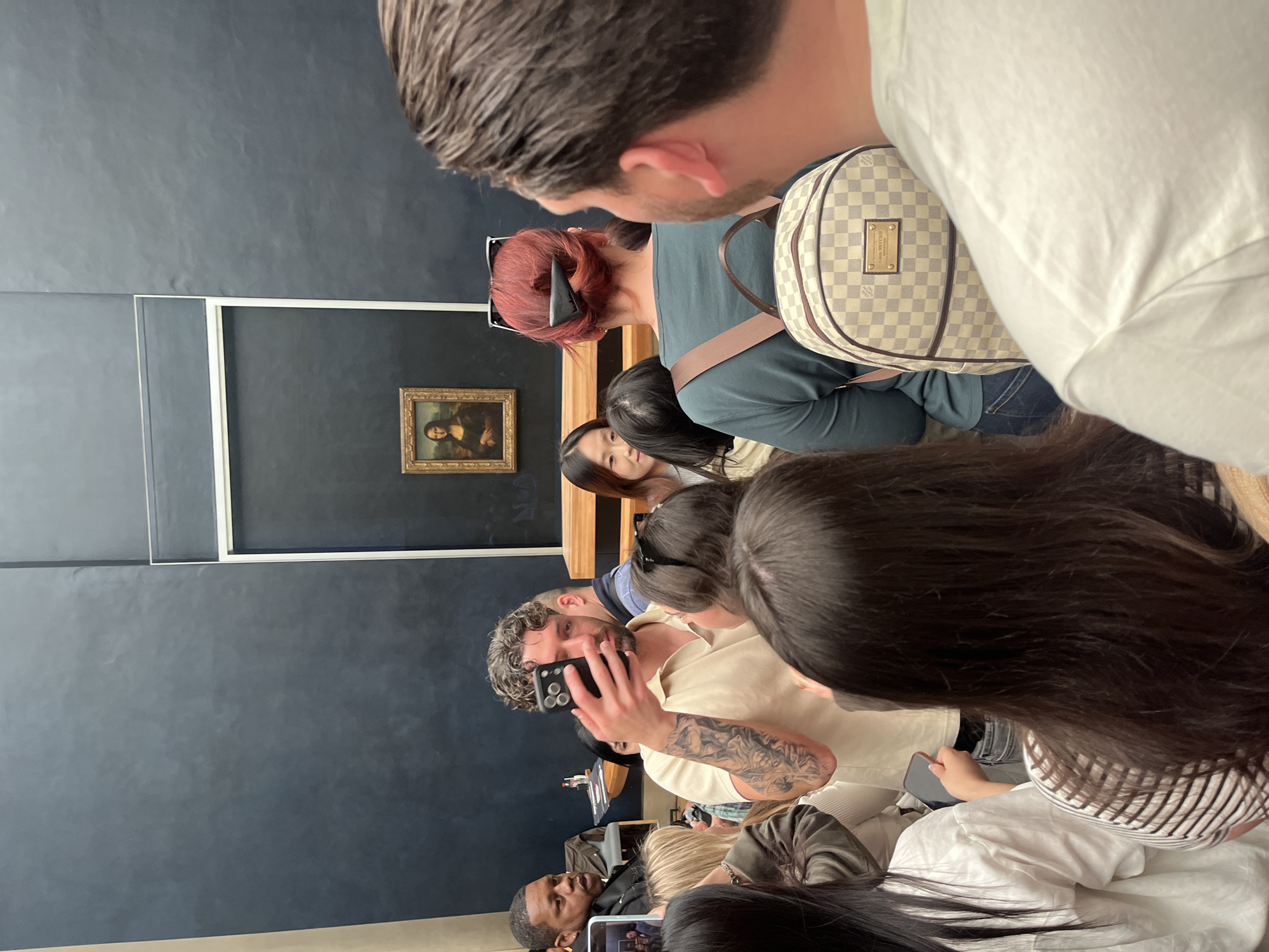 The image, rotated ninety degrees counter-clockwise, captures a bustling scene within an art museum, focusing on a crowd gathered before the renowned Mona Lisa painting. The famous artwork, encased in a golden frame, is prominently displayed against a dark, likely navy blue or charcoal gray, wall in the mid-ground. A white barrier or frame separates the viewers from the painting.
The foreground is dominated by the backs and profiles of several people. A man with brown hair and a white shirt is seen from behind, along with a woman with reddish-brown hair wearing sunglasses on her head. This woman is carrying a Louis Vuitton backpack featuring a checkered pattern, with "Louis Vuitton" and "Paris" visible on its label. Further into the crowd, a man with curly hair and a noticeable tattoo on his arm holds up a smartphone, seemingly taking a photograph or video of the Mona Lisa. Other visitors are also seen holding up their phones, indicating a common activity of documenting the experience.
The overall scene conveys the popular and often crowded nature of visiting such a significant cultural artifact. The setting, with its distinctive painting, dark walls, and large gathering of international visitors, definitively places the location as the Louvre Museum in Paris, France. The image captures the moment of engagement between spectators and one of the world's most iconic masterpieces.