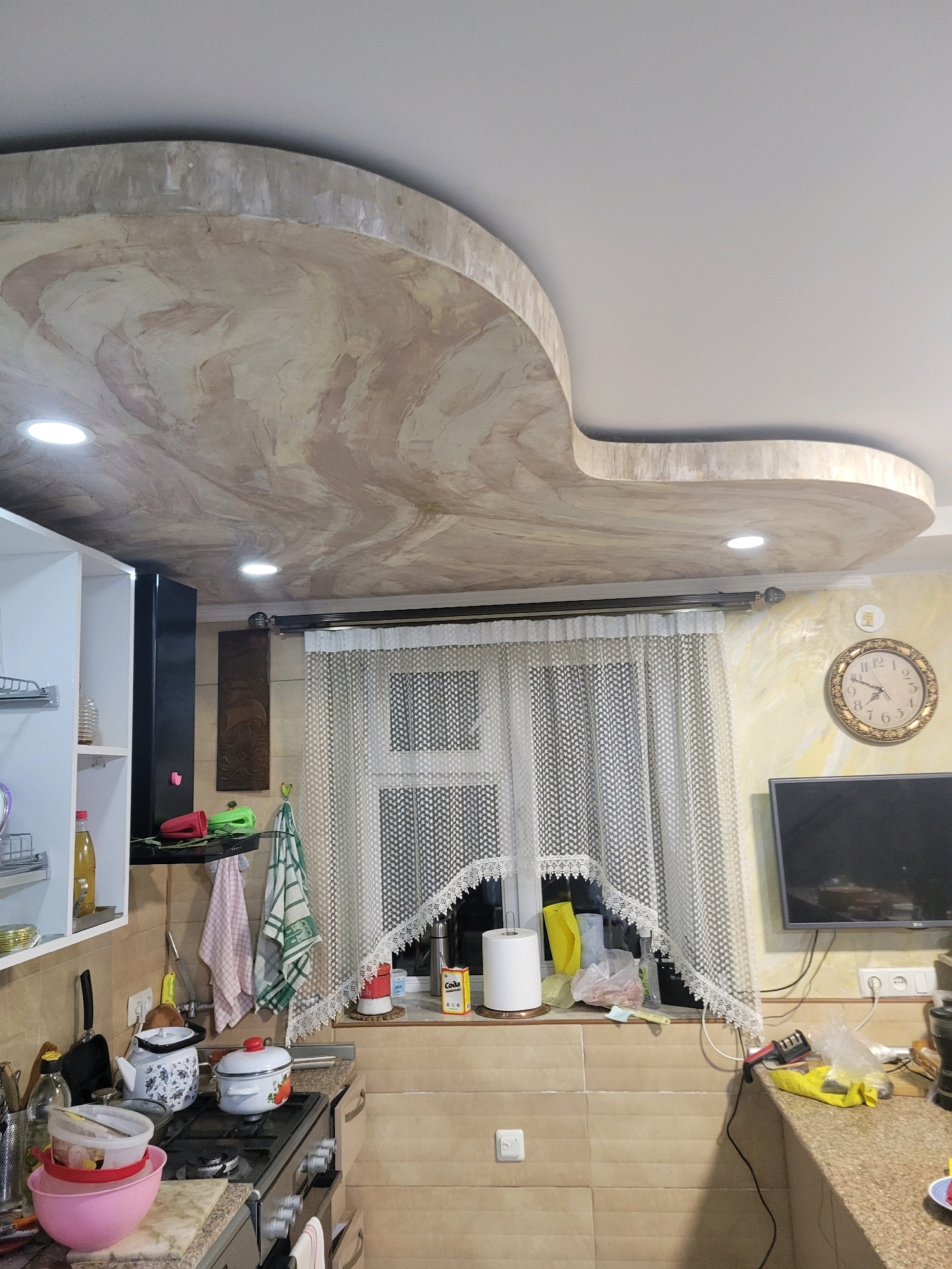 The image depicts an interior scene, likely a kitchen, with a distinctive curved ceiling design featuring a marbled pattern. The ceiling has recessed lighting. In the background, a window is covered by a white, lacy curtain. To the right of the window, a wall-mounted television is visible, and above it, a circular clock with a gold frame is hung. The kitchen counter space below the window is cluttered with various items, including a roll of paper towels, plastic bags, and cleaning supplies. A stovetop with pots and pans is present on the counter, along with kitchen utensils and bowls. A white cabinet unit is on the left side of the image. The walls are tiled with a light beige material. The overall impression is of a lived-in kitchen. There are no people visible in the image. The location is identified as Sachkhere, Georgia.