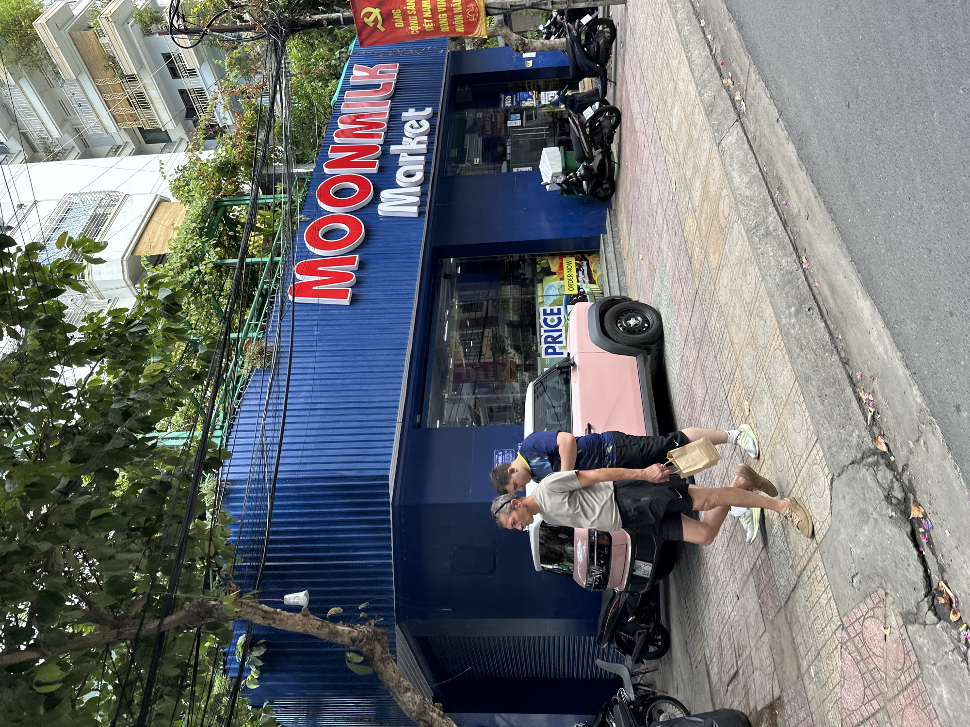 The image captures a street scene in Nha Trang, Vietnam, featuring a blue building with a prominent "MOONMILK Market" sign in red and white lettering. In front of the market, two men are walking on the sidewalk, one carrying a paper bag. A small, light pink vehicle is parked near the entrance of the market. Several motorcycles are parked along the street. The building's facade is characterized by vertical blue corrugated metal. The surrounding environment includes trees, electrical wires, and a multi-story building in the background. A red banner with Vietnamese text and imagery is visible on the right side of the frame, partially obscured by a tree. The sidewalk is paved with patterned tiles, and the street is asphalt.