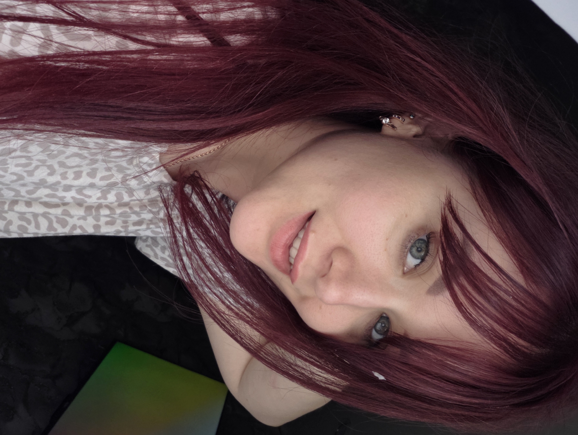 A close-up, high-angle selfie shows a young woman with long, dark red hair and pale skin with freckles. She has light blue-green eyes, light-colored eyebrows, and a small nose. Her lips are painted a light pink. She is wearing a light-colored t-shirt with a grey, abstract pattern and a thin gold necklace. She has two earrings in her left ear, one small stud and one hoop with a small gem. Her left arm is bent and resting behind her head, and her right hand is visible near her ear. The background is dark, with a hint of a dark, textured surface on the right, possibly a table, and a corner of a reflective object with green and pink hues. The location is Chaikovsky, Russia.