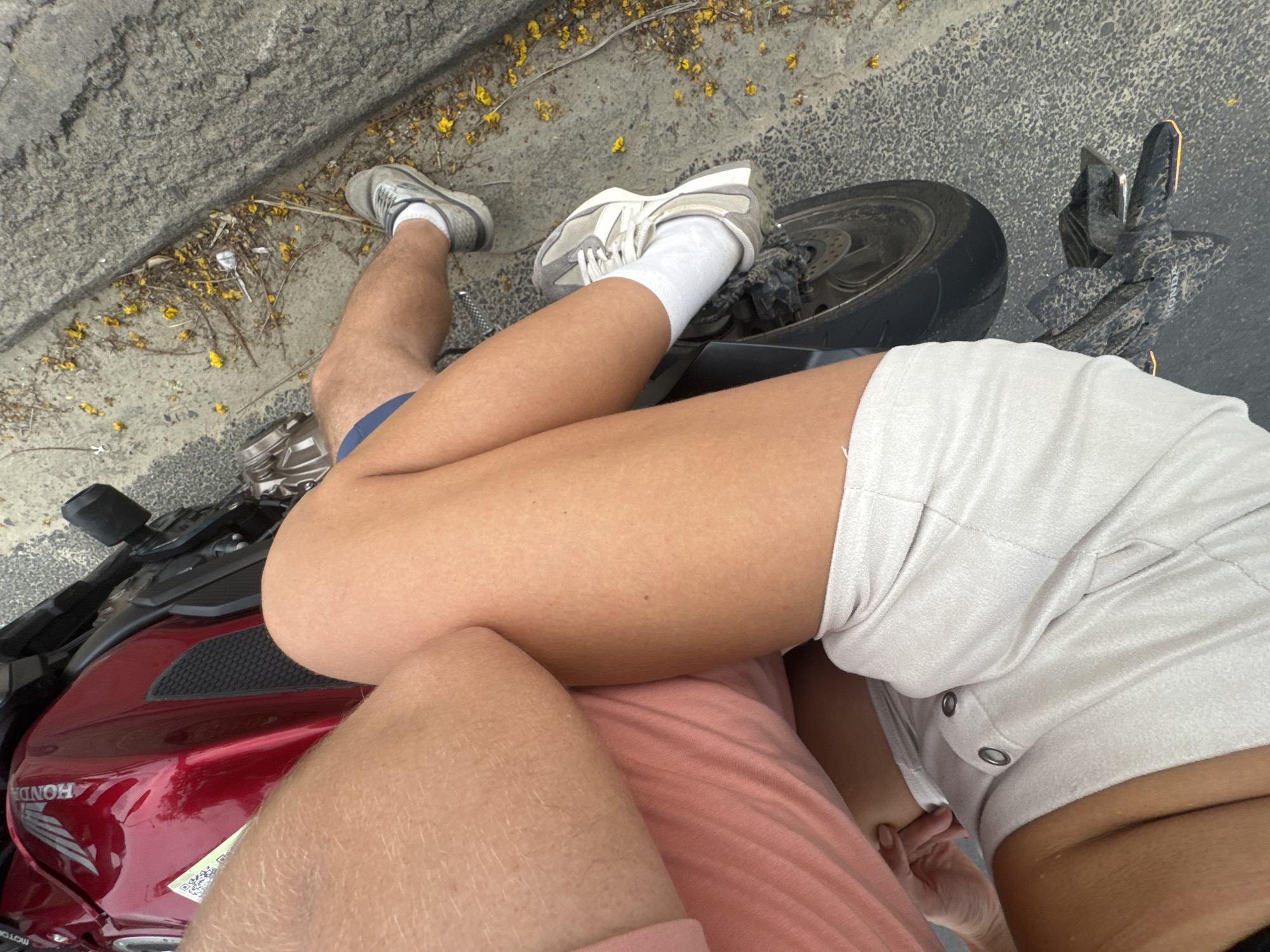 A high-angle close-up shot shows a person seated on a parked red Honda motorcycle. The motorcycle is positioned near a curb with scattered yellow blossoms. The person wears a pink t-shirt, light-colored shorts, white socks, and grey sneakers. Another pair of dark-colored shoes are placed next to the rear wheel of the motorcycle. The specific location in Đà Nẵng, Vietnam, cannot be determined from the image alone.