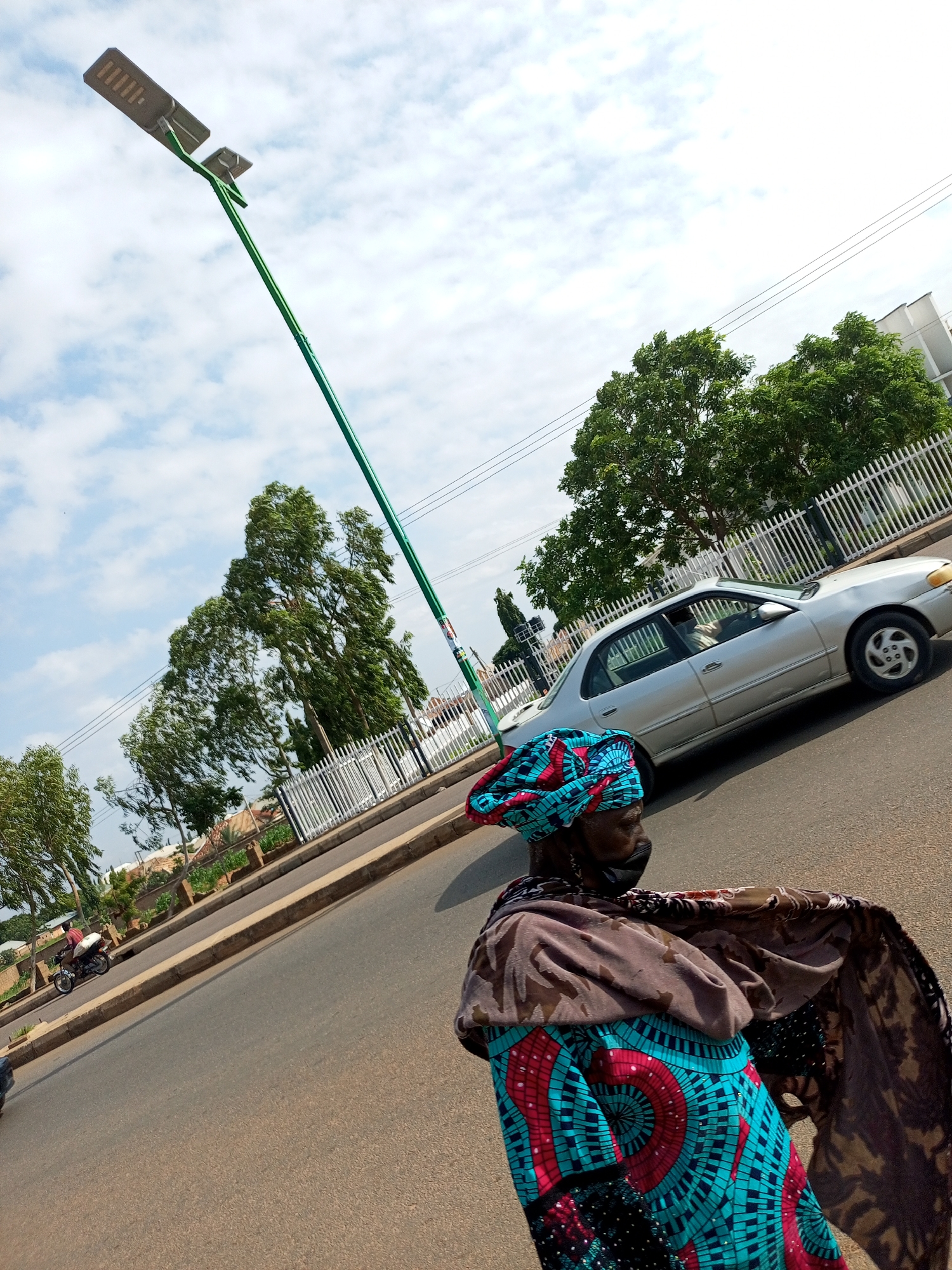 The image depicts a street scene in Tumfure, Nigeria, on a cloudy day. In the foreground, a woman walks along the road, wearing a vibrant blue and red headwrap and a patterned dress, with a dark face mask covering her nose and mouth. She is draped in a brown patterned shawl. To her right, a silver sedan is driving on the road. In the background, a tall, green lamppost with two solar panels extends upwards into the sky. Trees with lush green foliage line the side of the road, behind which a white picket fence is visible. Further in the distance, more trees and some buildings can be seen. A motorcycle with a rider is also present on the far left side of the image, on the side of the road. The overall scene suggests an ordinary day in a Nigerian urban environment.
