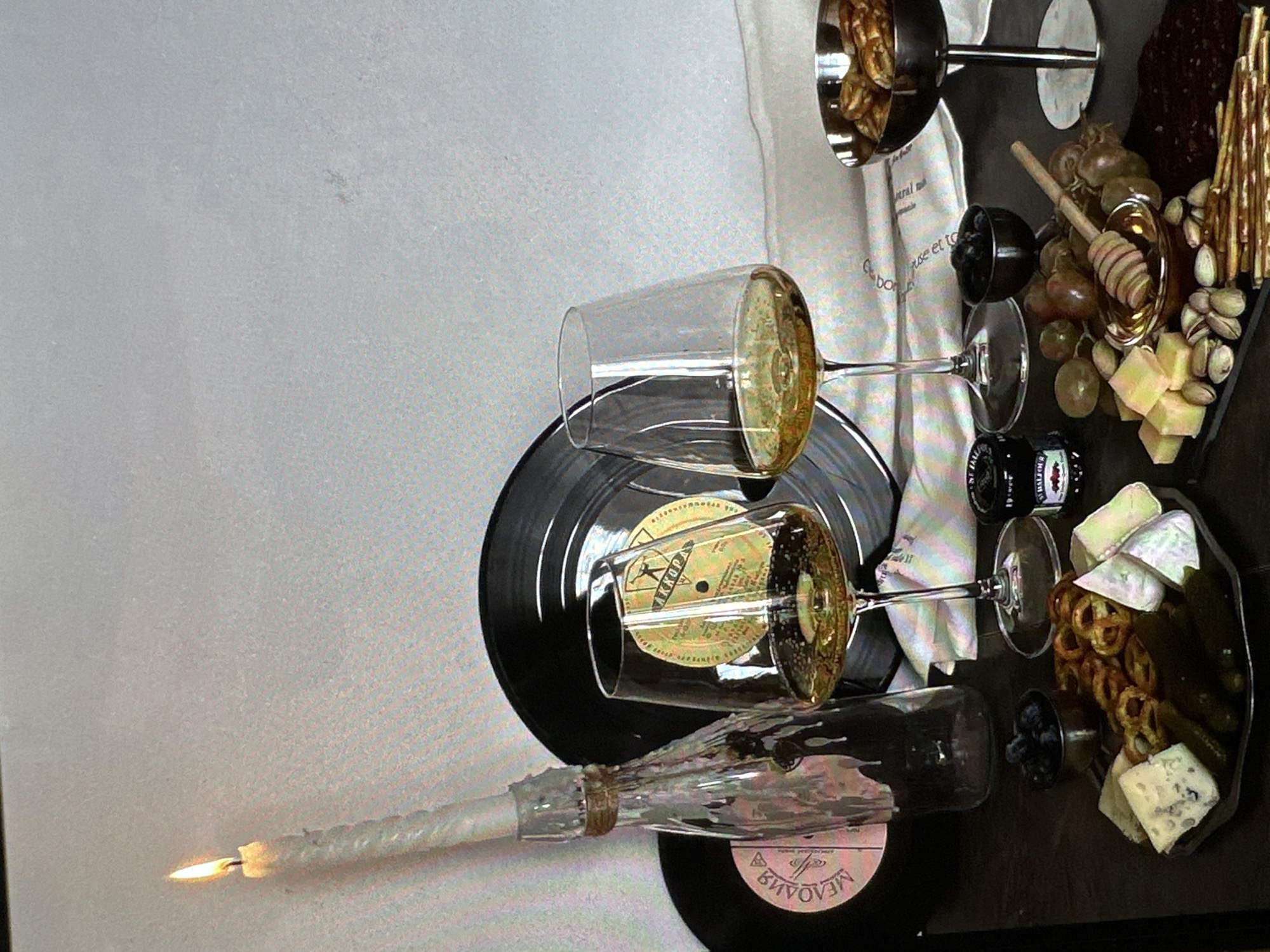The image presents a still life arrangement on a table, presumably indoors. There are two vinyl records; one is black with a tan label bearing non-Latin script, and the other is black with a pink label also with non-Latin script. Two partially filled wine glasses rest on the records. A long, burning candle lays horizontally across the lower record. A black serving tray holds a charcuterie assortment, including grapes, cheeses, pretzels, pickles, and breadsticks. Two small metal bowls contain blueberries and a condiment. Behind the serving tray, there is a white cloth with text in a foreign language and a metal serving dish containing what appear to be breadsticks. The background is a plain wall. There's no indication of Yerevan, Armenia.