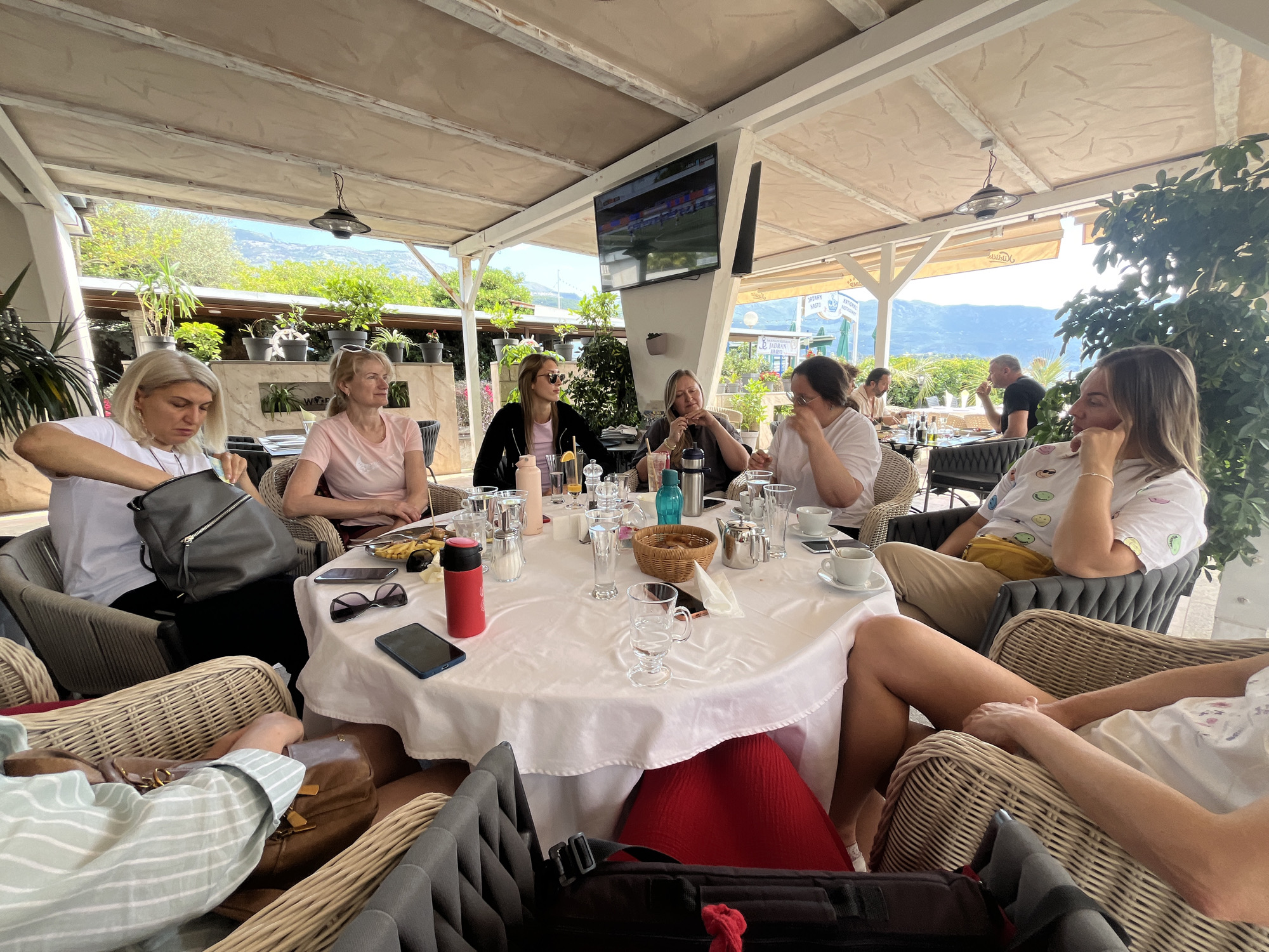 A group of eight women are seated around a large, round table with a white tablecloth at an outdoor restaurant. The location is in Budva, Montenegro, as indicated by the surrounding landscape. The restaurant is covered by a partial roof structure. There is a TV visible overhead on a support beam. The table is filled with various items including glasses, a teapot, a basket, drinks, and personal items such as a phone and sunglasses. The women are engaged in conversation and appear to be enjoying their meal or drinks. The chairs are made of woven material. Plants and trees are visible in the background, adding to the outdoor ambiance. Other restaurant patrons are seated in the background.