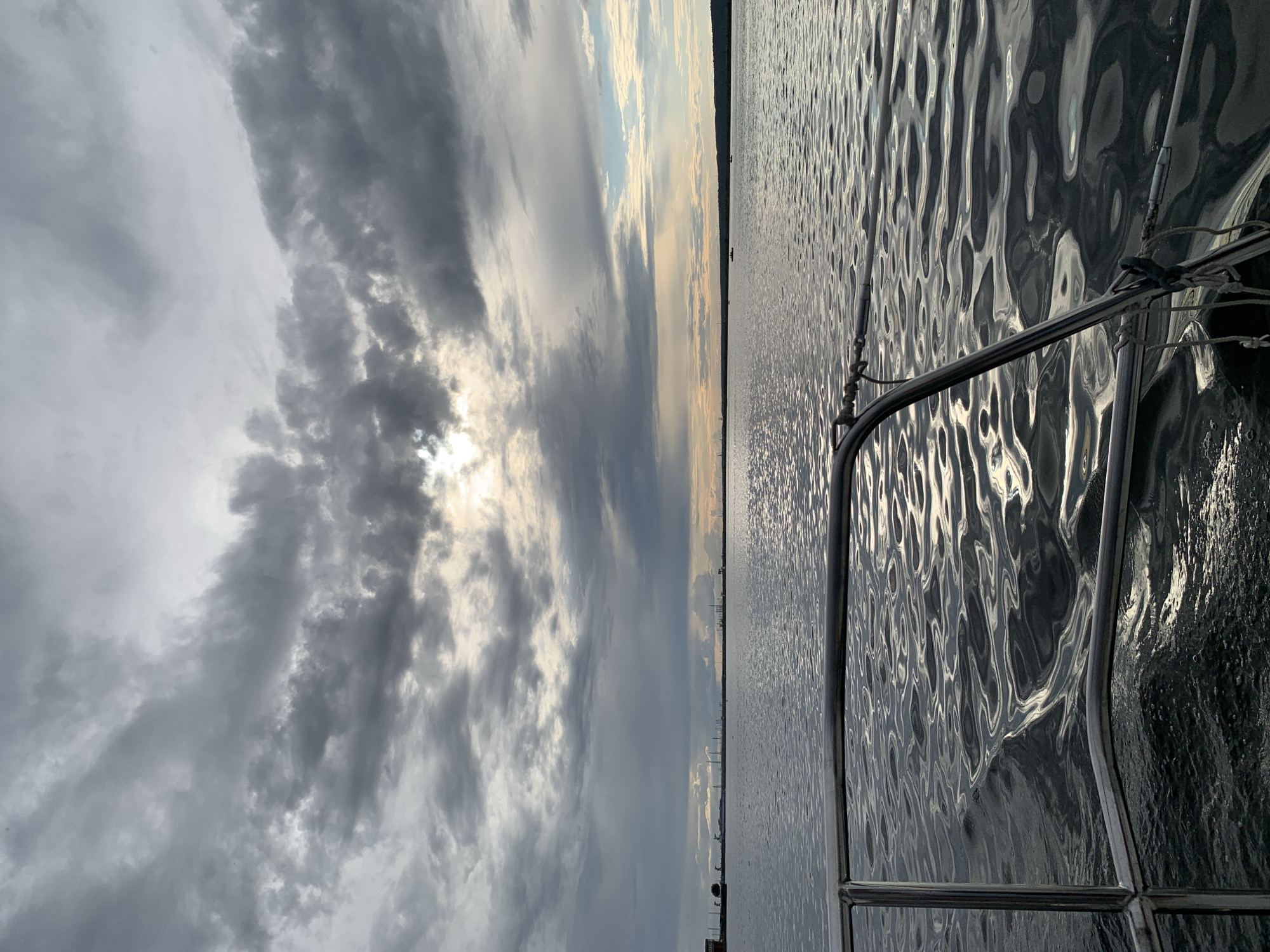 The image depicts a view from a boat on a body of water, likely a river or reservoir, with a cloudy sky overhead. In the distance, a silhouetted industrial skyline is visible along the horizon, characteristic of the industrial port area of Chaikovsky, Russia. The water's surface is rippled, reflecting the muted light filtering through the clouds. The foreground shows the metal railing of the boat. The overall scene suggests a calm, overcast day.
