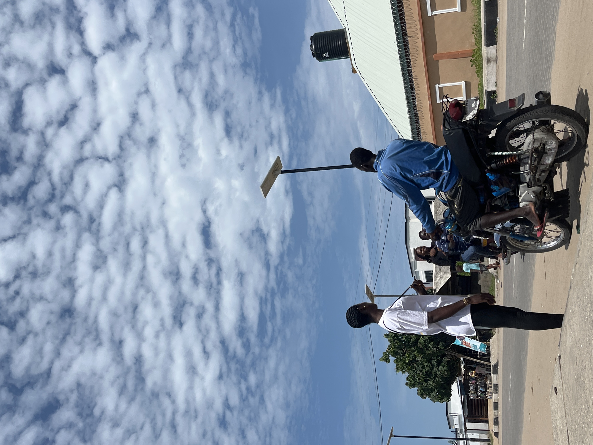 The image captures an urban scene in Badagry, Nigeria, characterized by a partly cloudy sky above a street. A man in a white shirt and black pants stands on the sidewalk. A man is seated on a motorcycle. A metal pole extends upward from the motorcycle, with a flat rectangular object attached to the end of the pole. Several other people stand near the motorcycle. Buildings with metal roofs and windows are visible along the street. A water tank sits on a building. A tree is located close to the sidewalk.