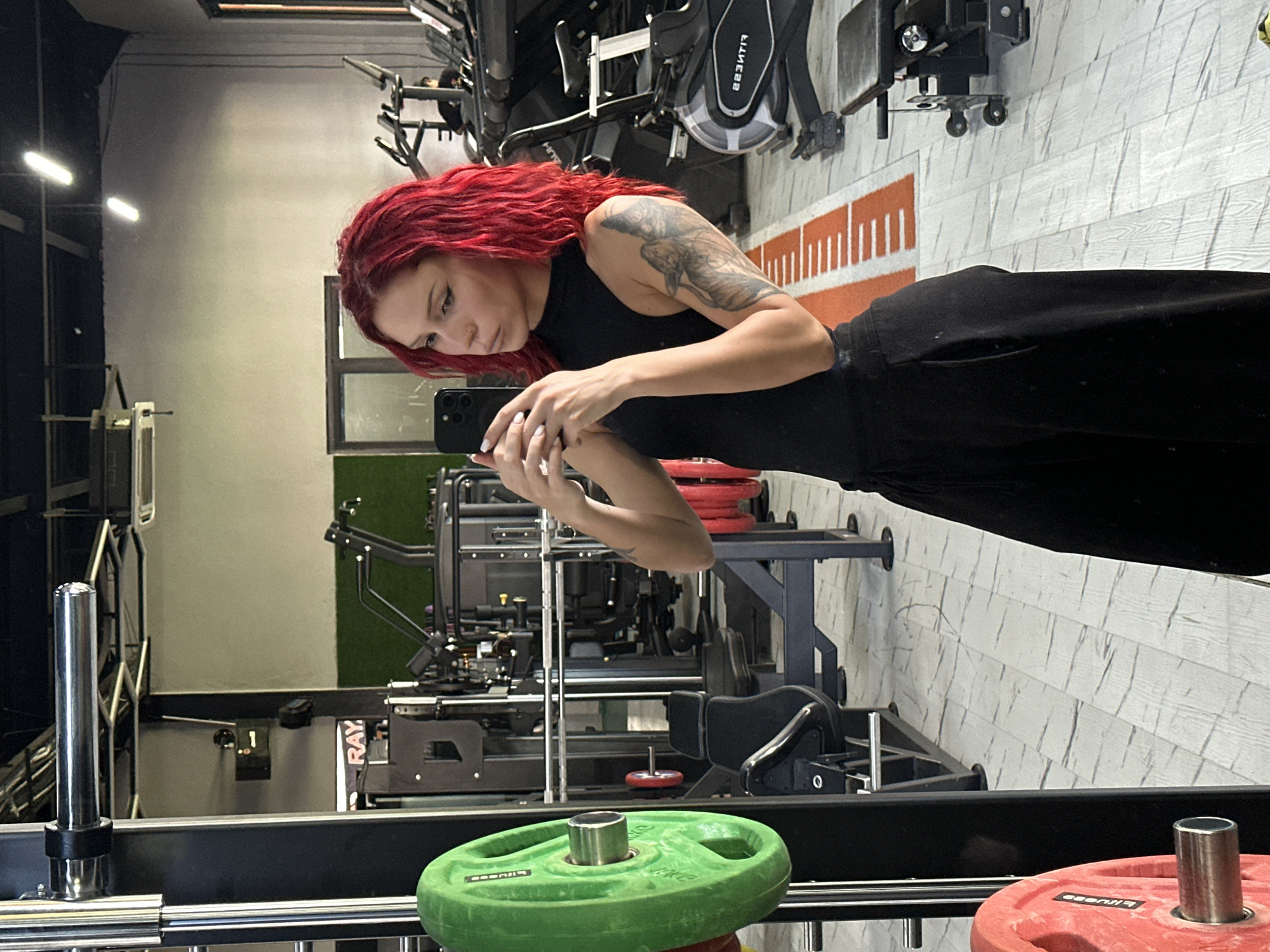 The image displays an indoor fitness center or gym located in North Kuta, Indonesia. A female individual with bright red, curly hair and a tattoo depicting a feline-like creature on her left upper arm is positioned in the center, taking a mirror selfie with a dark-colored smartphone that features a triple camera array. She wears a black sleeveless high-neck top and black wide-leg trousers; her fingernails are painted white, and she has a nose ring.
The gym floor is covered with light gray and white patterned tiles. A prominent orange and white track-like marking, resembling a measurement scale, extends across the floor from the mid-left to the mid-right. To the left, a section of a black Smith machine or weight rack is visible, with a large, circular green weight plate (marked "10KG" and "BITUGOD") attached to a chrome barbell sleeve. A red weight plate is partially visible below the green one.
In the background, various pieces of gym equipment are present, including multiple treadmills (one displaying mirrored text resembling "223ITJIT" on its console), an exercise bike, and black upholstered weight benches with silver frames. A section of artificial green turf is visible in the far background on the right, suggesting a functional training area. The walls are painted light gray, and overhead fluorescent strip lighting illuminates the space. A partially obscured red sign reading "RAY" is visible on the wall behind the equipment on the left, along with a black wall-mounted box and a small black bag hanging below it. An air conditioning unit is also visible on the ceiling.